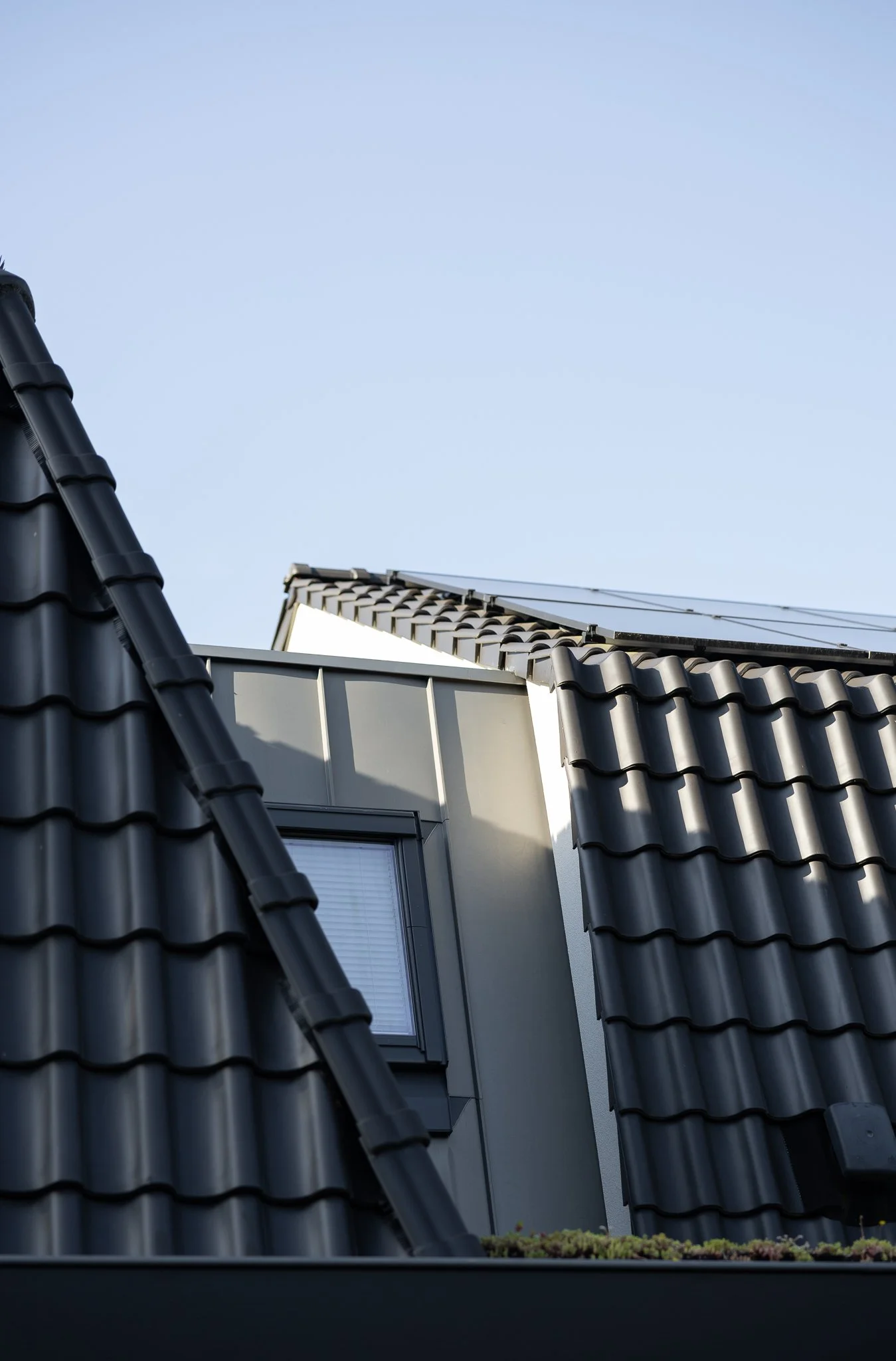 Dark tiled roof with dormer window, metal cladding and solar panels against a blue sky