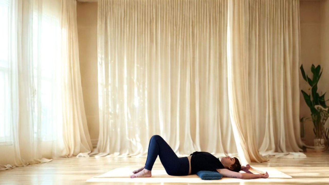 une femme allongée sur le sol un couverture en dessous des cotes, les genoux pliés, les pieds à plats sur le sol, des rideaux en fond . elle est apaisée, les yeux fermés