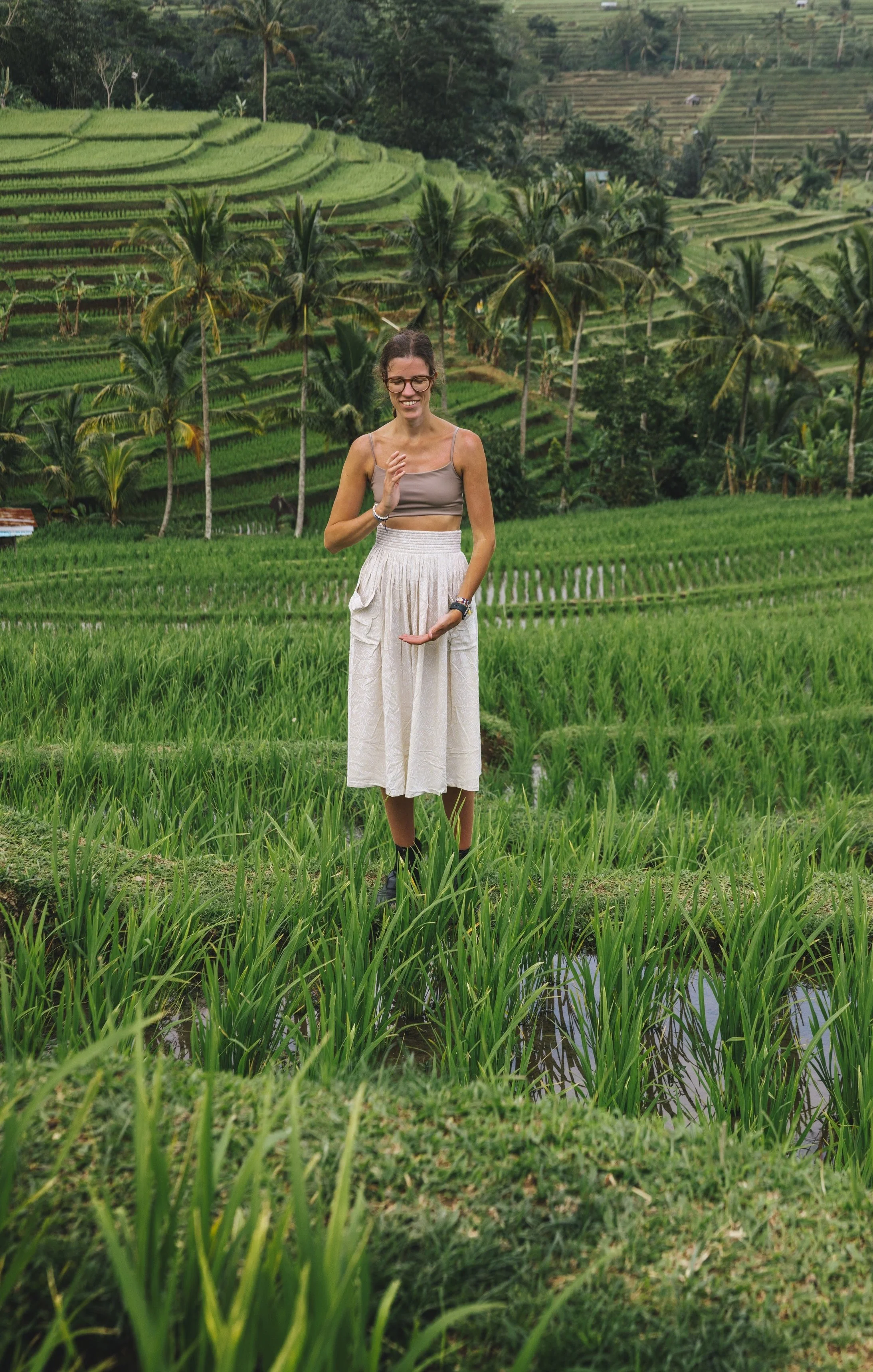 Eine lachende Frau steht auf einer Plantage mit grünen Reisfeldern und Palmen im Hintergrund. Linda Zimmermeier hält die Energie im Herzraum für das Herz Qi Gong.