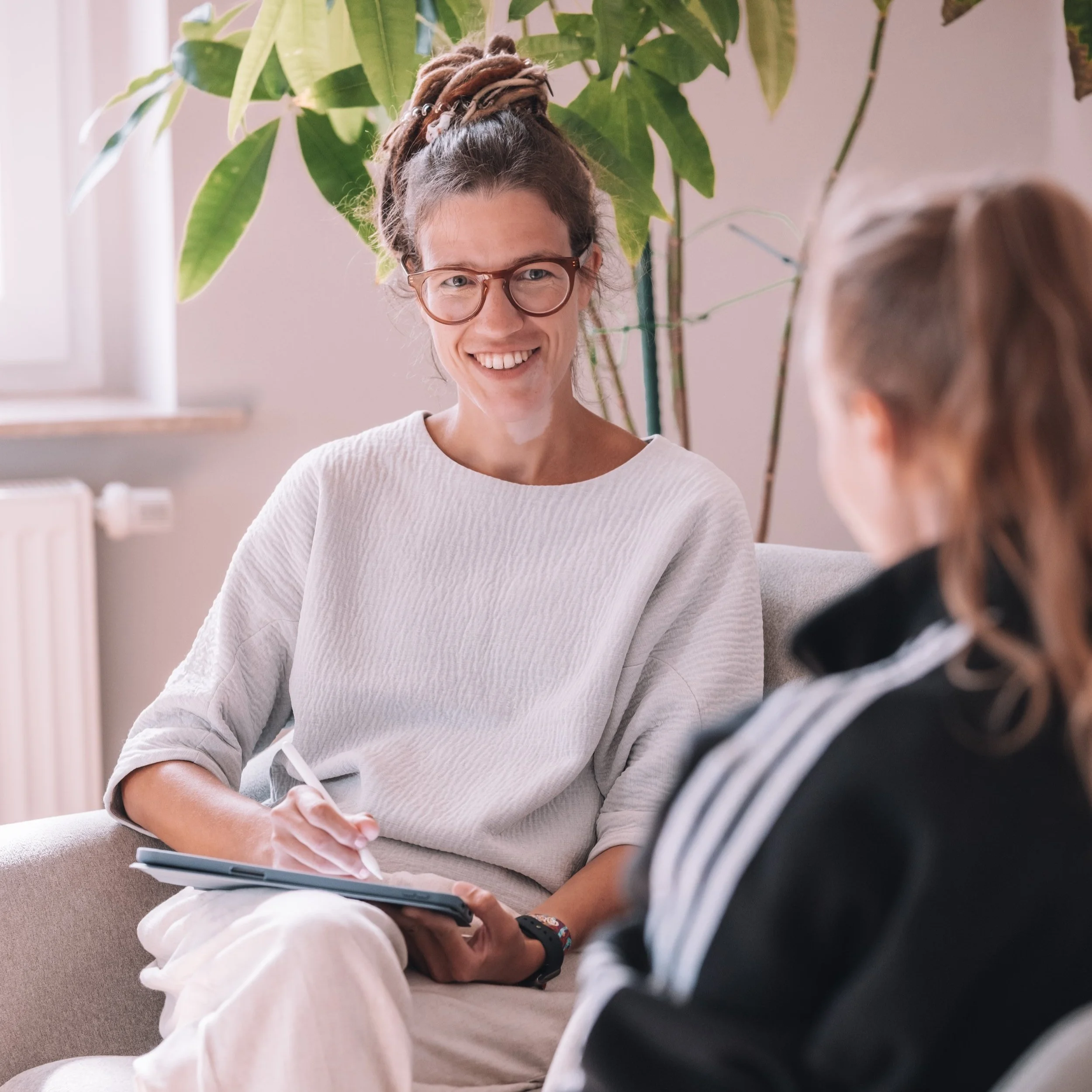 Zwei Frauen im Gespräch in einem hellen Raum mit einer großen Pflanze im Hintergrund. Linda Zimmermeier führt ein ausführliches Anamnesegespräch mit einer Patientin durch.