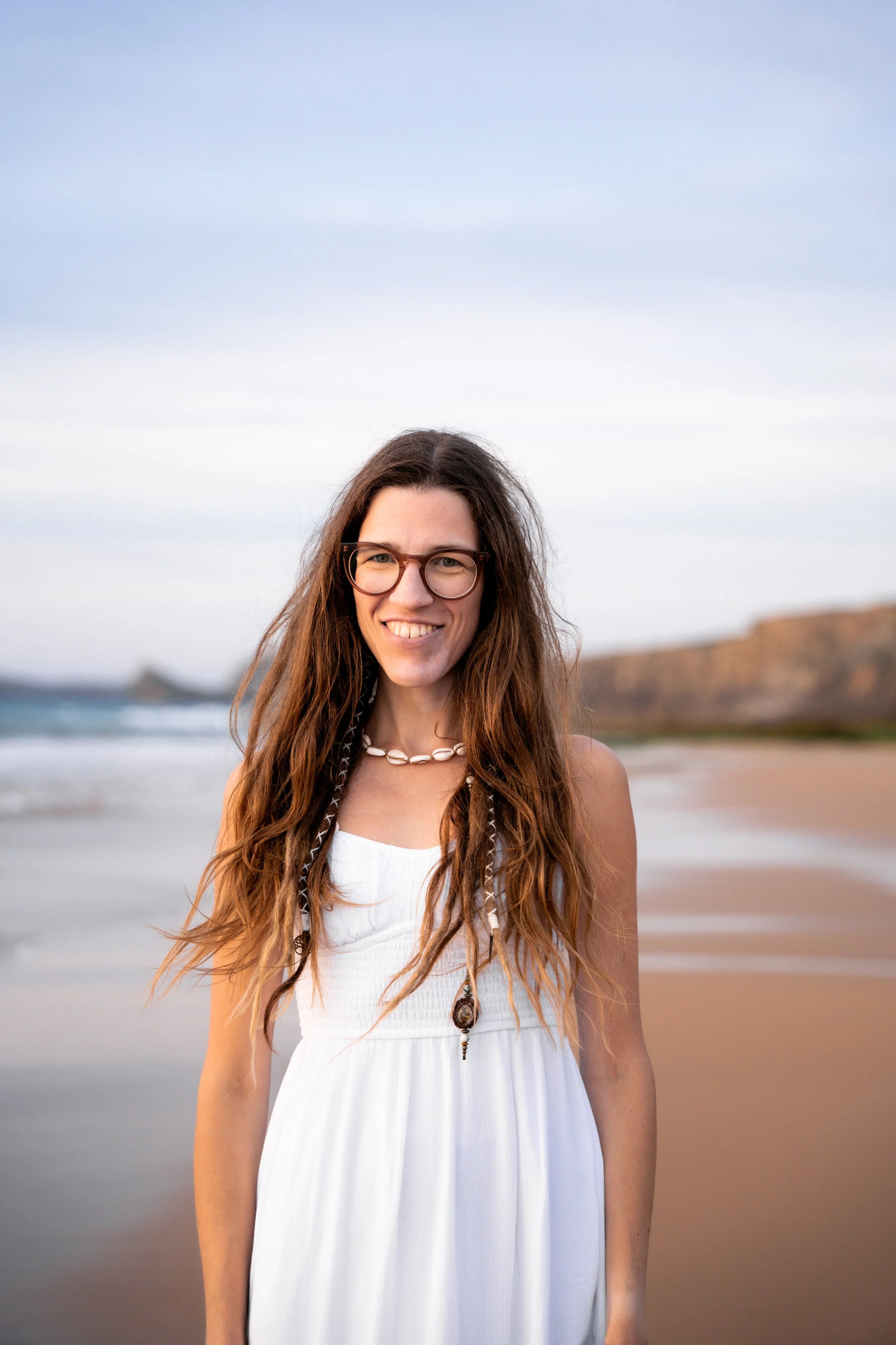 Die Heilpraktikerin für Physiotherapie Linda Zimmermeier ist am Strand, trägt Brille und weißes Kleid, lächelt in die Kamera, im Hintergrund Felsen und Wasser, bewölkter Himmel. Das Bild strahlt Leichtigkeit und Ruhe aus.