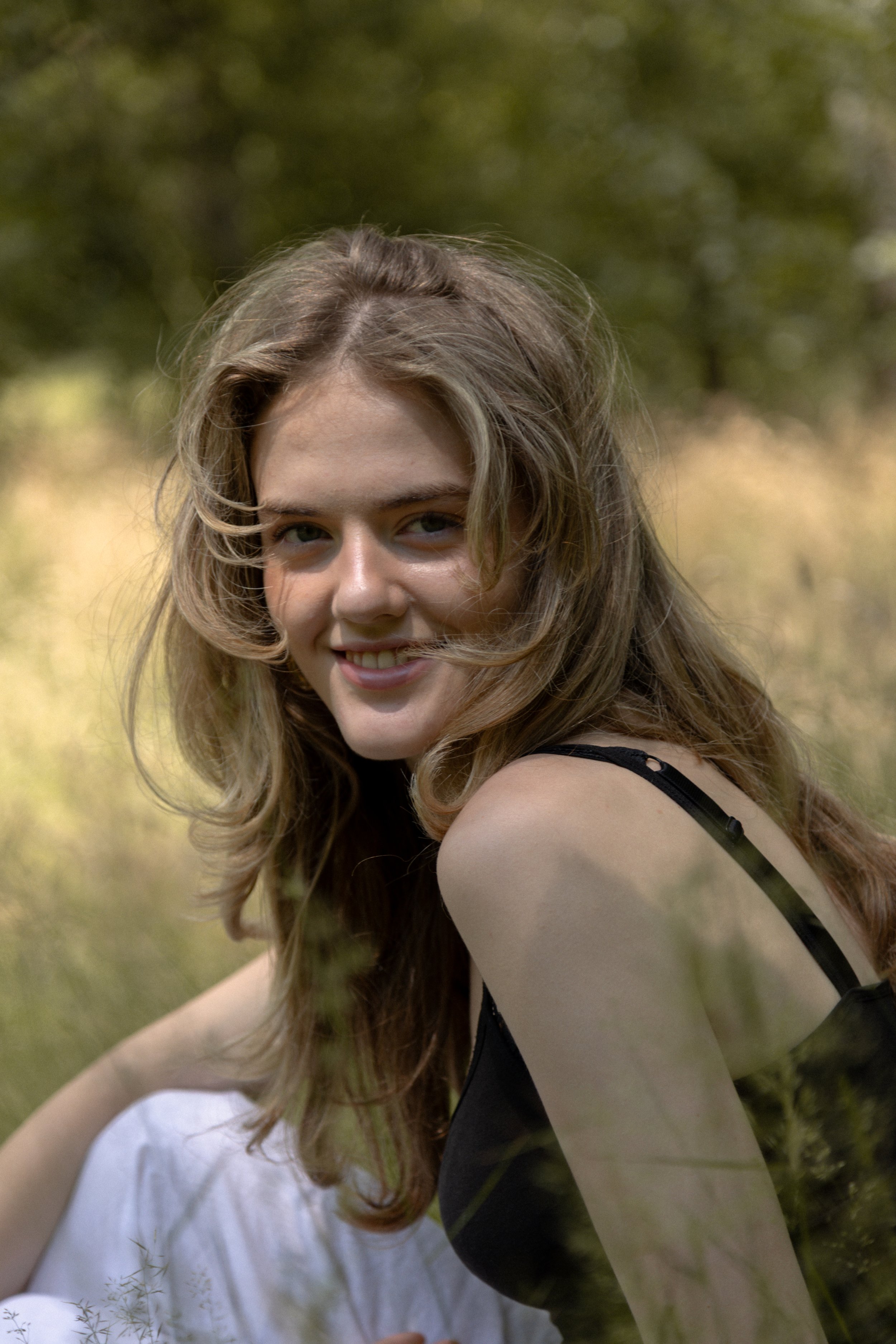A young woman with long, wavy brown hair smiling outdoors in a green, natural setting with tall grass and trees.