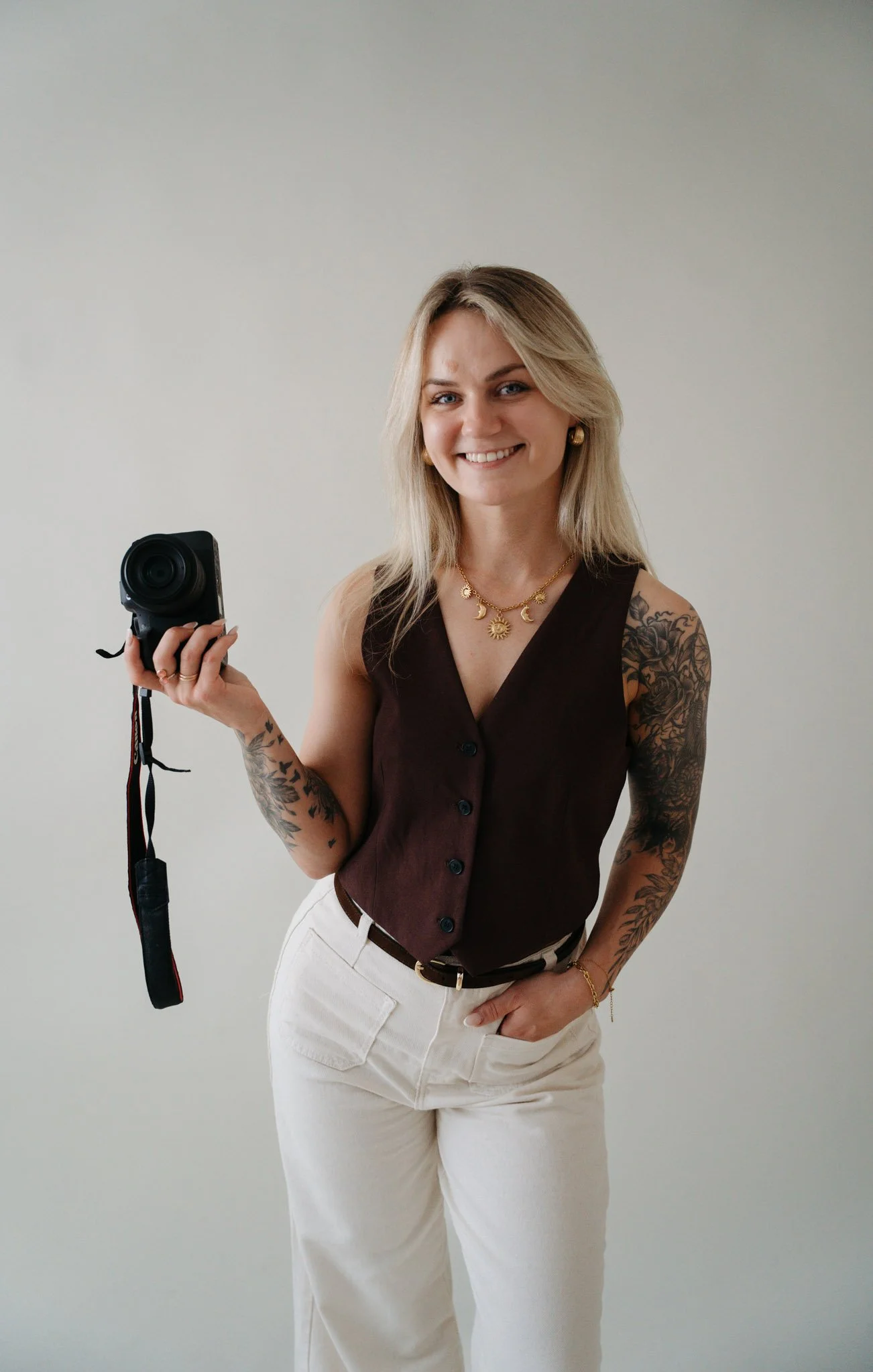 Smiling woman with blonde hair, tattoos on her arms, wearing a sleeveless dark top and light-colored pants, holding a camera in her right hand, standing against a plain light background.