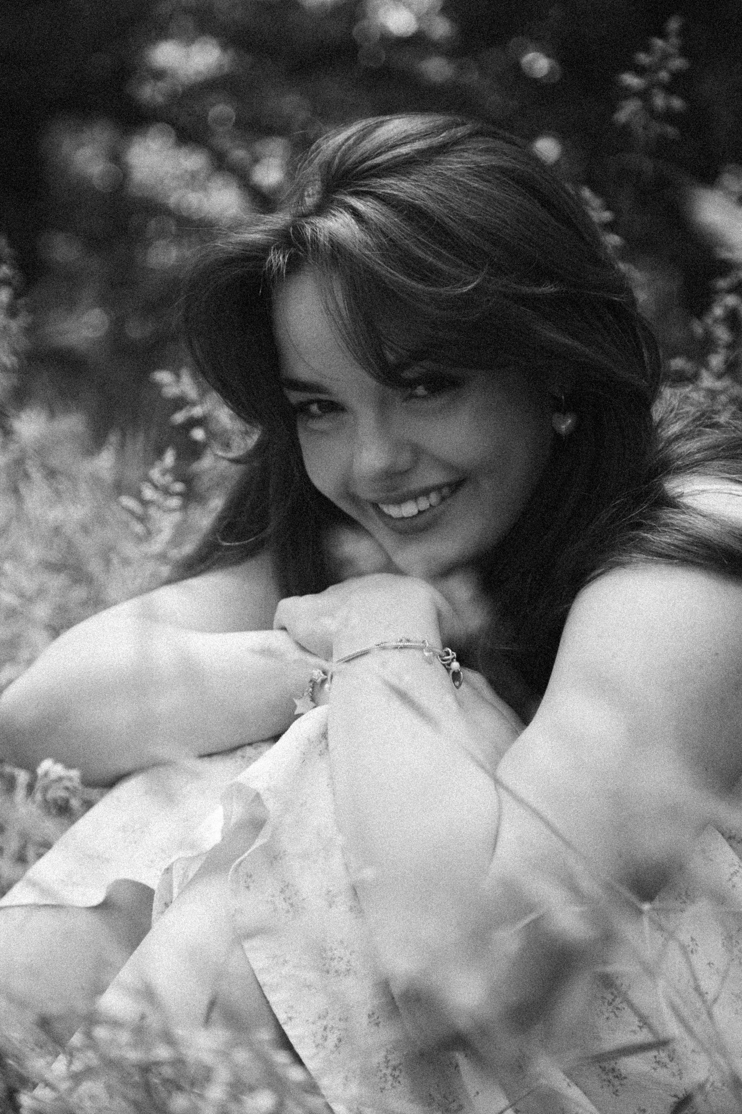 Black and white photograph of a smiling woman with styled hair, wearing earrings and bracelets, resting her arm on her knee, outdoors with blurred foliage in the background.