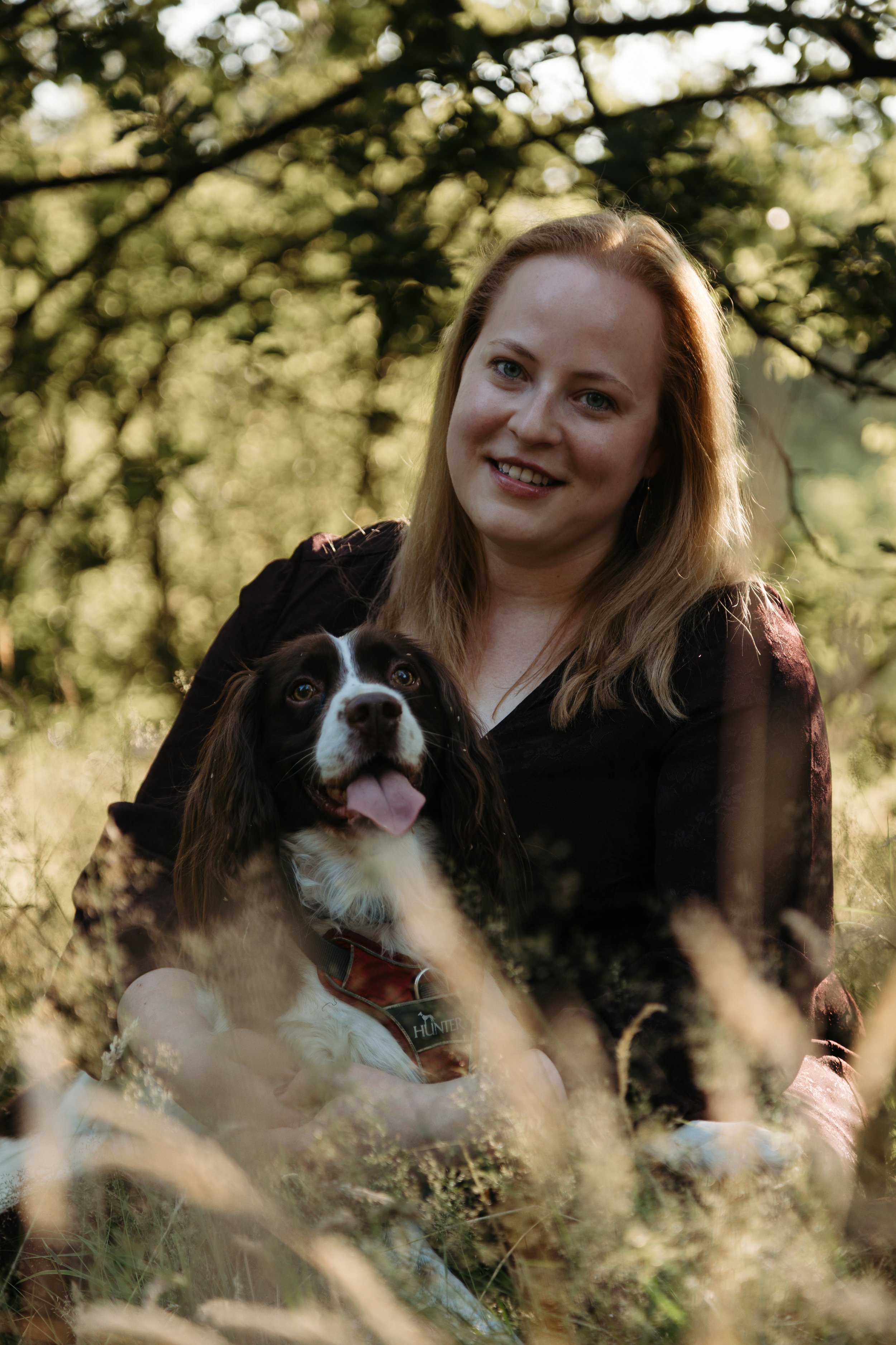 A woman with blonde hair smiling while holding a black and white dog that has a tongue out, sitting outdoors among grass and trees.