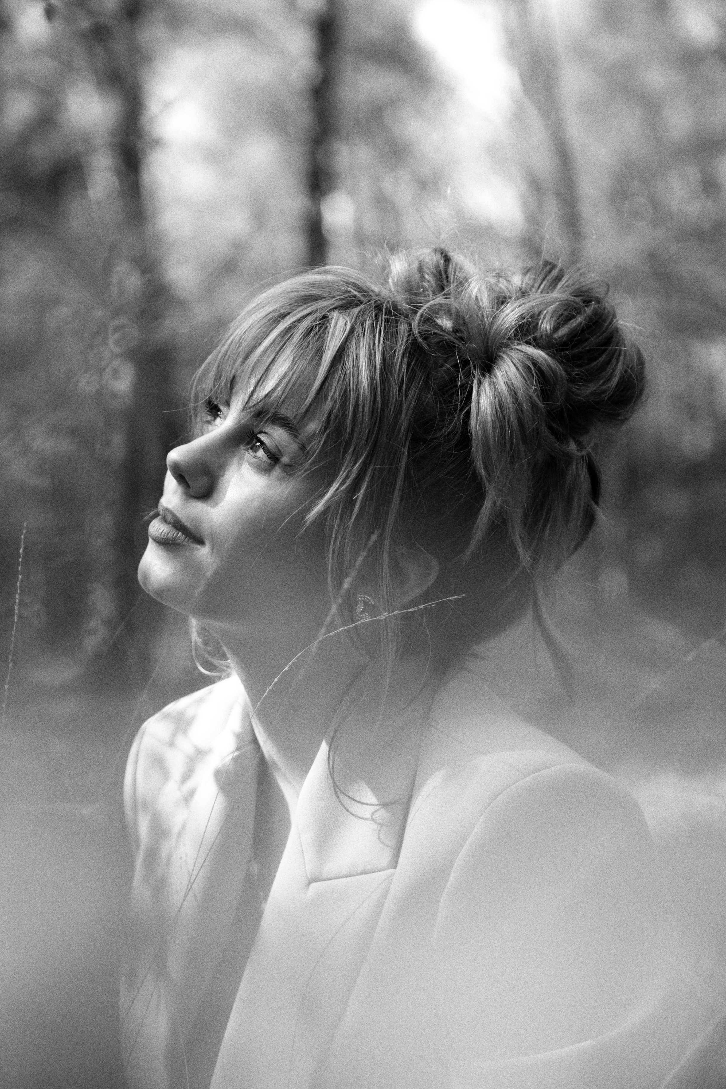A black and white portrait of a woman with curly hair, looking thoughtfully into the distance outdoors, with a blurred background of trees.