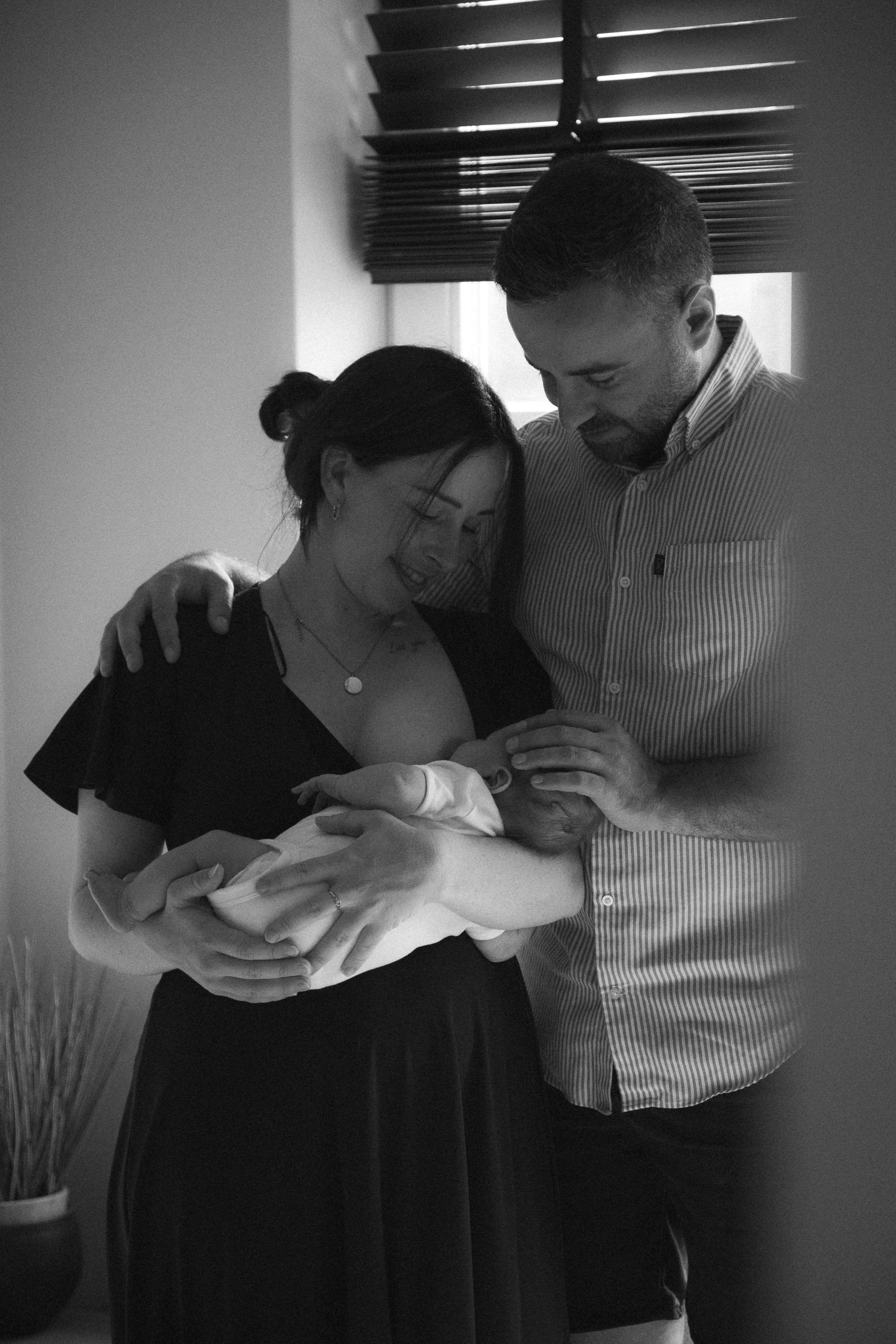 Black and white photo of a couple holding their newborn baby. The woman is smiling and looking at the baby, while the man is gently touching the baby's head. They are standing near a window with blinds.