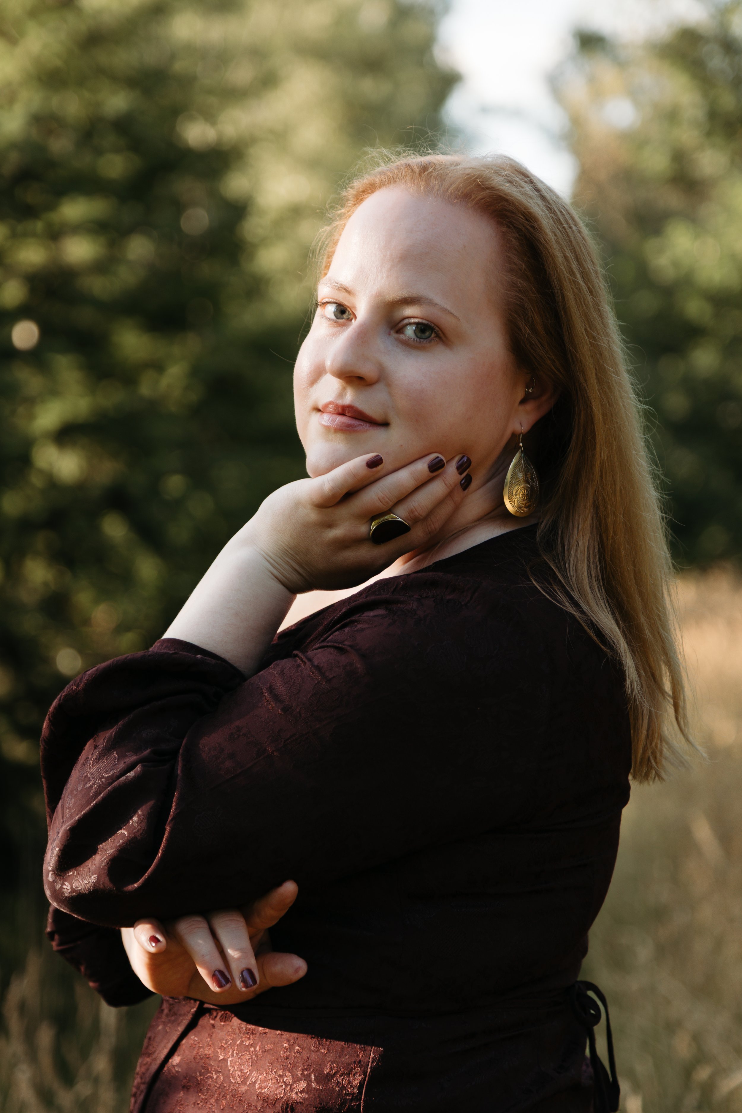 A woman with light skin and reddish hair outdoors, touching her chin with her hand, wearing earrings, a large ring, and a dark-colored top. She is looking at the camera with a slight smile. Background is blurred greenery.