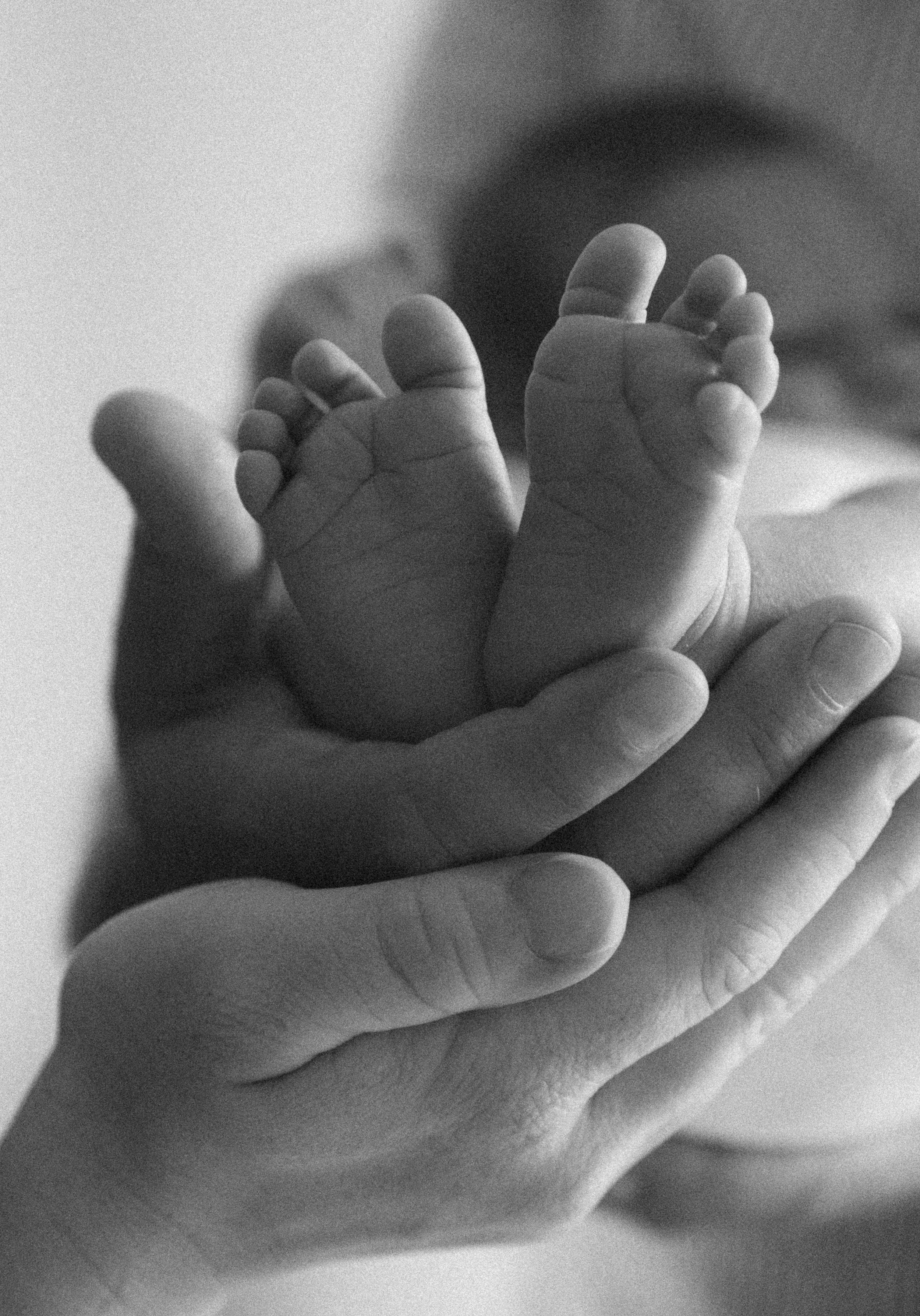 A close-up black and white photo of a small baby's hand grasping an adult's finger, with a blurred baby in the background.