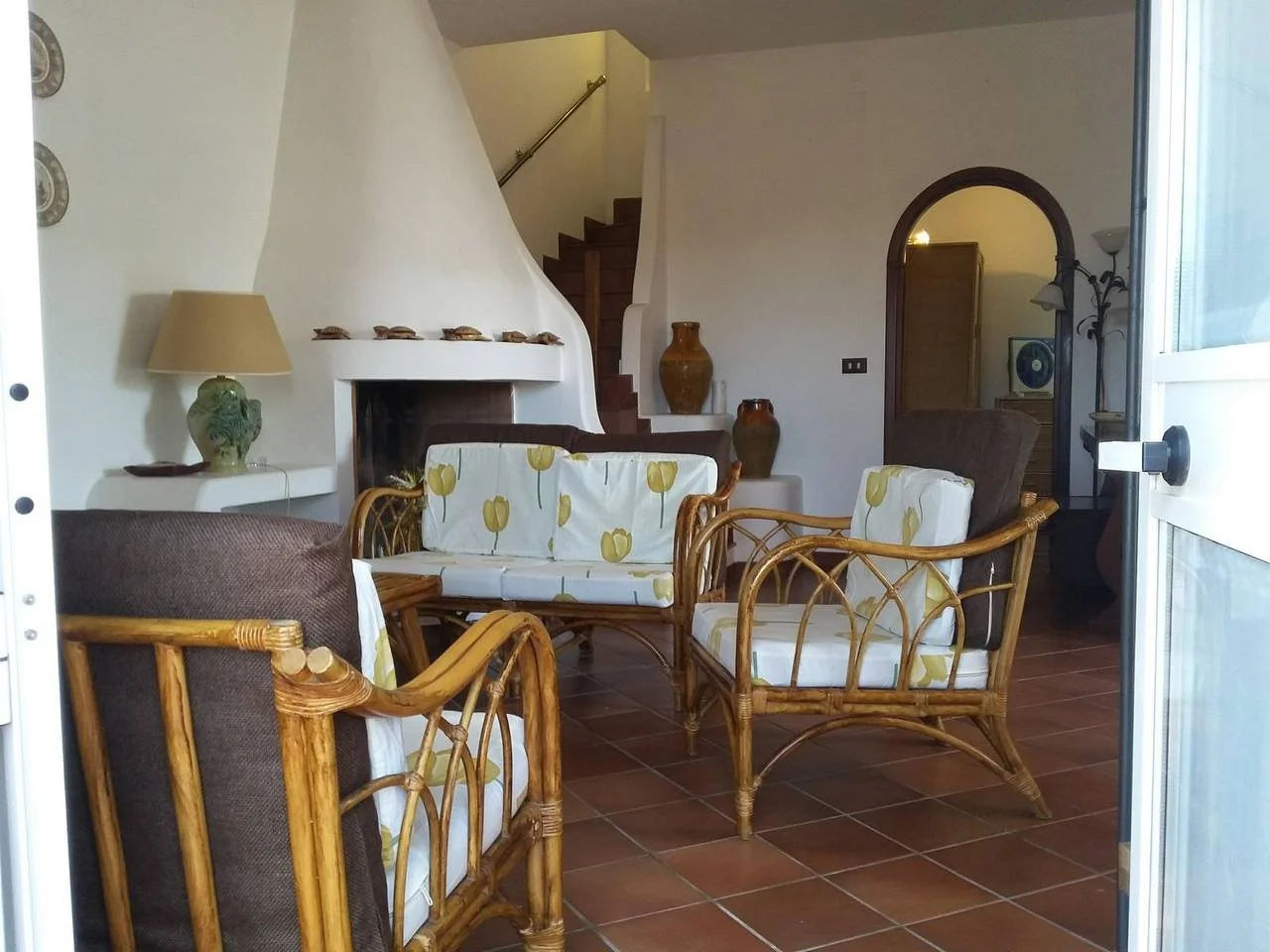 Soggiorno con divani in vimini, camino bianco, tappeti e vasi di terracotta, ingresso con scala e porta a arco.