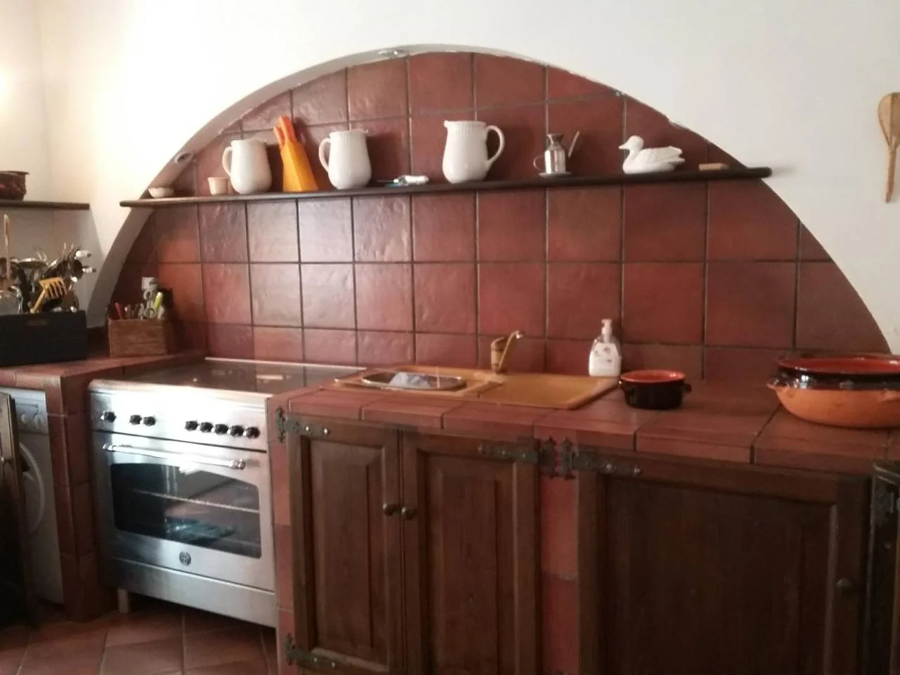 Cucina con parete in mattoni rossi, elementi di cucina in legno, forno a gas, ripiano con mattarello e sapone liquido, decorazioni in porcellana bianca e un cigno di ceramica