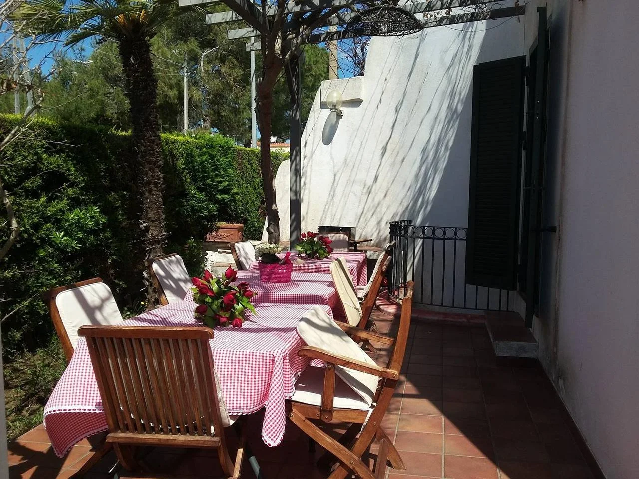 Tavolo all'aperto con tovaglia a quadretti rossi e bianchi, decorato con bouquet di fiori rossi e una rosa bianca, su una terrazza soleggiata circondata da piante e cespugli