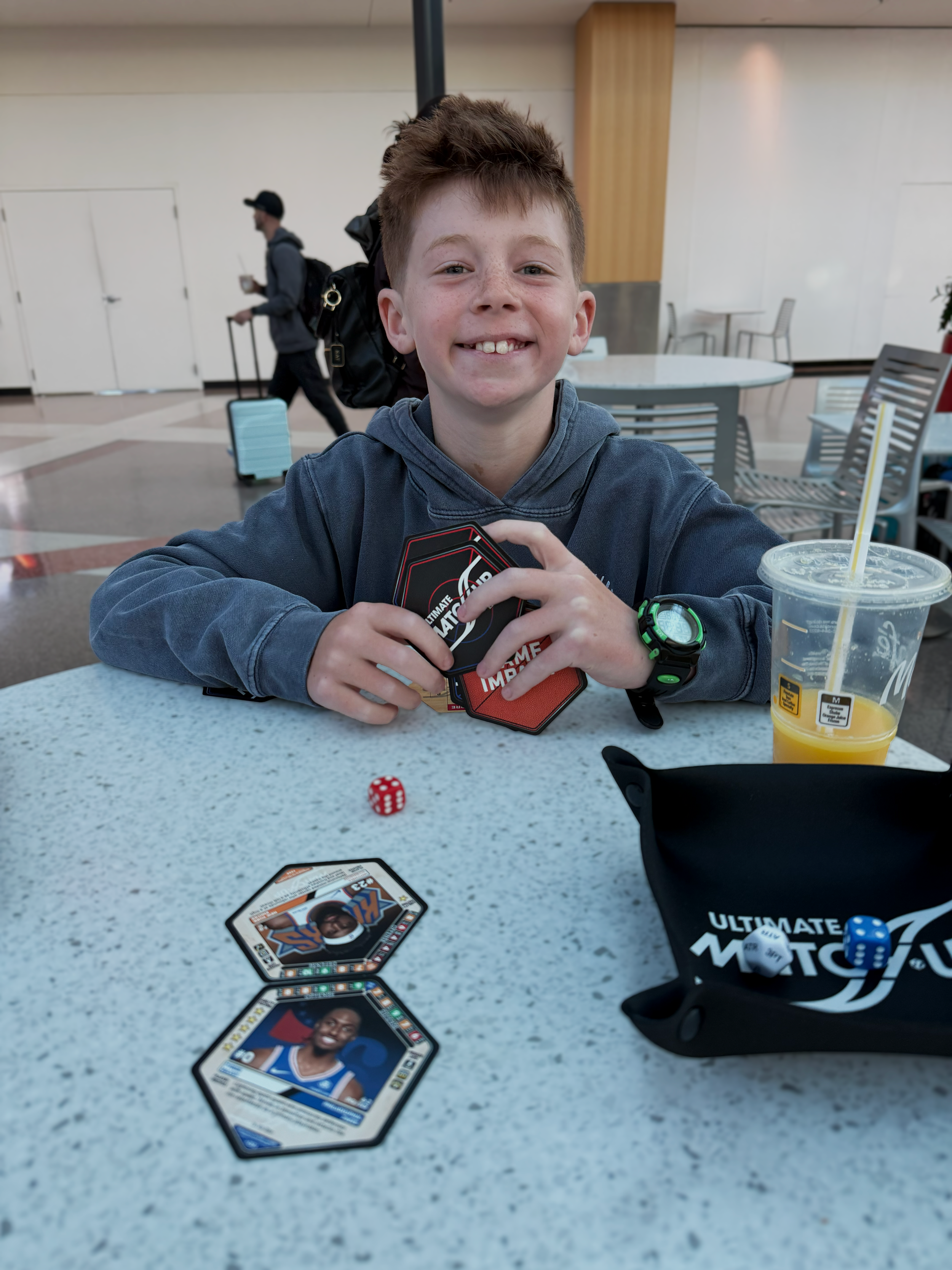 A young boy with short multifaceted hair smiling and holding cards at a table in an airport, with various playing cards, a die, a drink, and a black bag on the table. In the background, a man is walking with a backpack and a beverage.