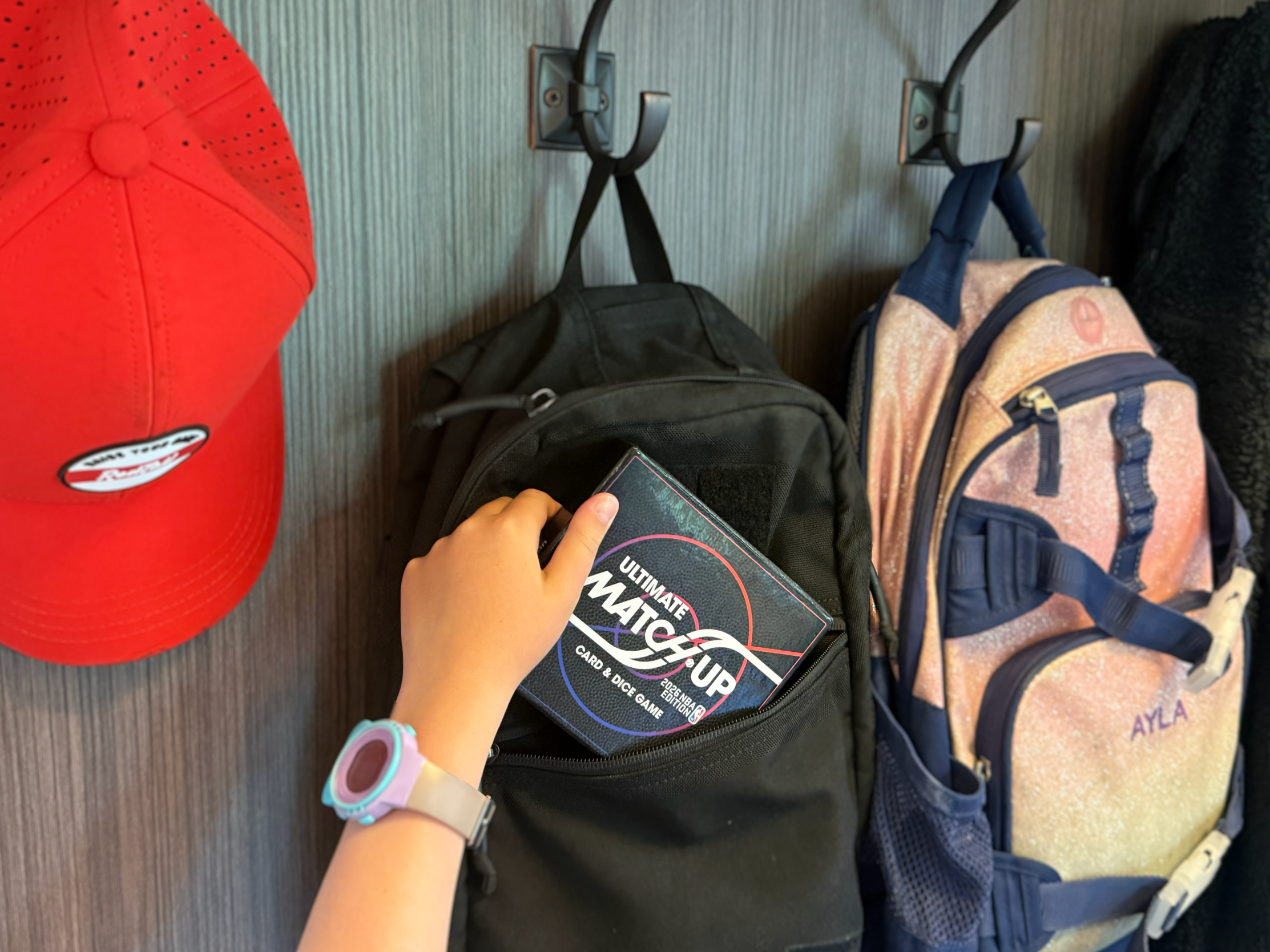 Person holding a card game box titled "Ultimate Match-Up" near backpacks and a red baseball cap on the ground.