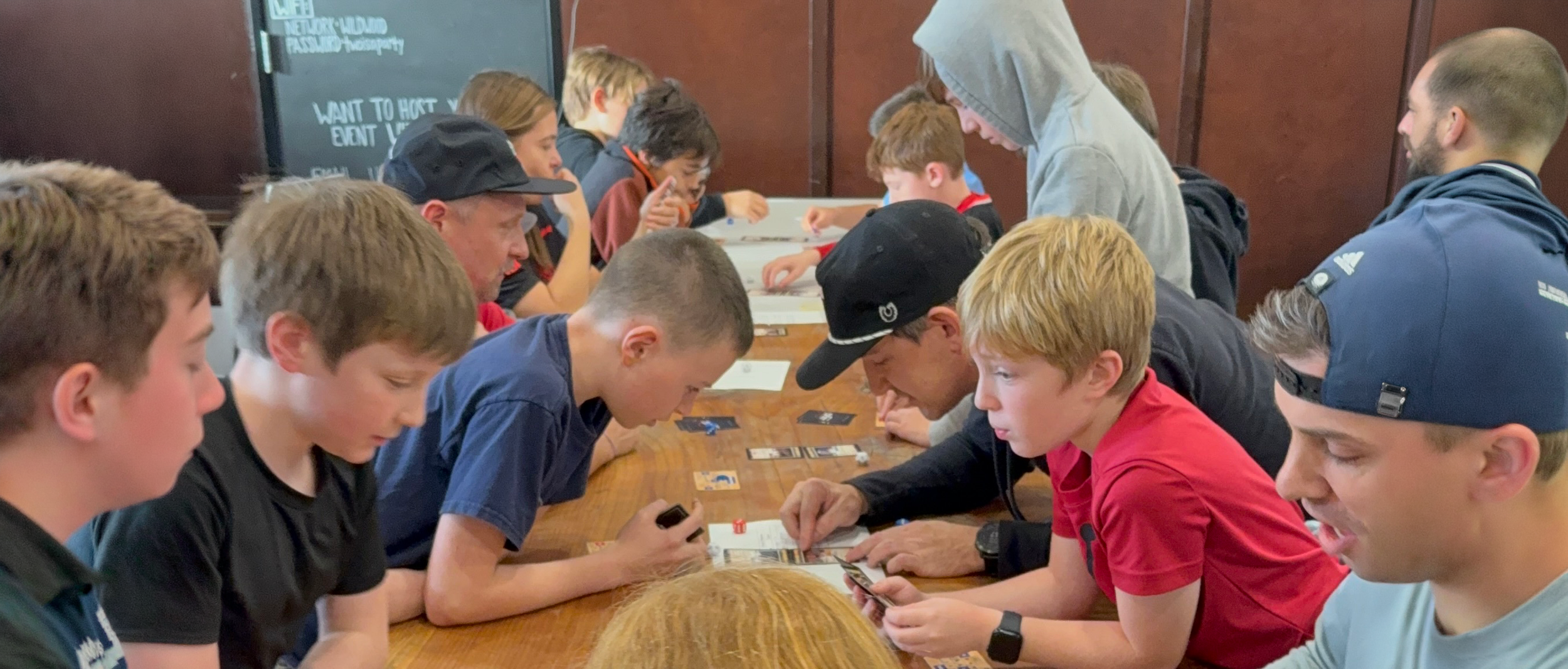 Group of young boys participating in a sports card game tournament around a wooden table in recreational setting
