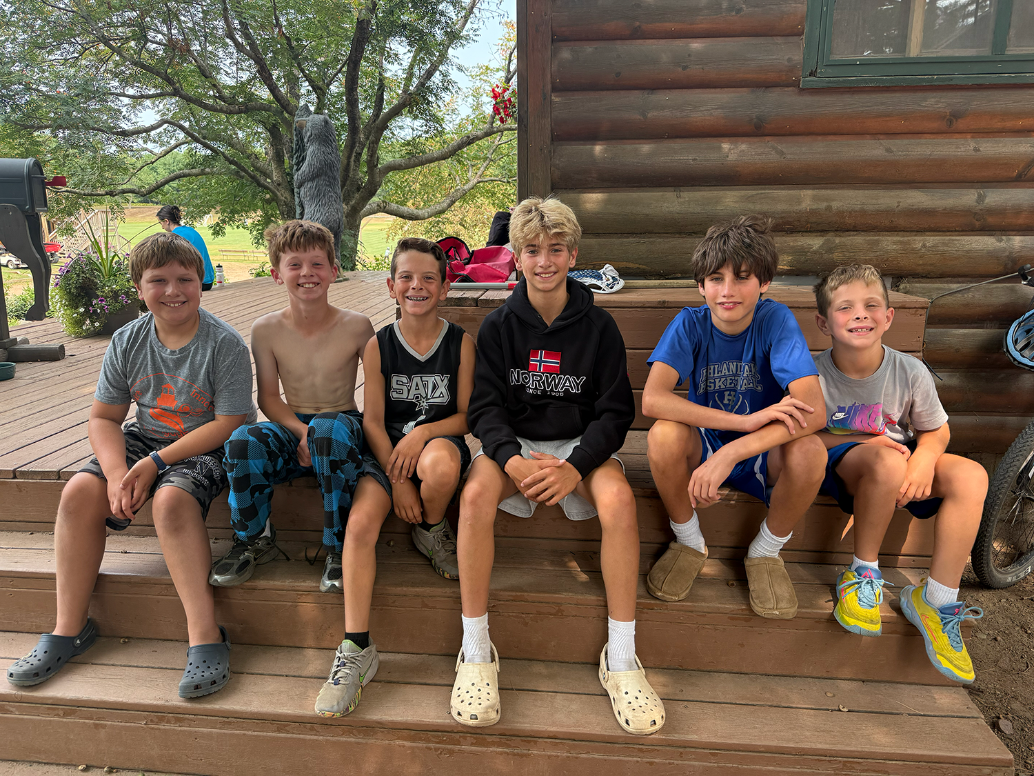 Six boys sitting on the porch of a wooden cabin, smiling at the camera, with trees and a woman in the background.