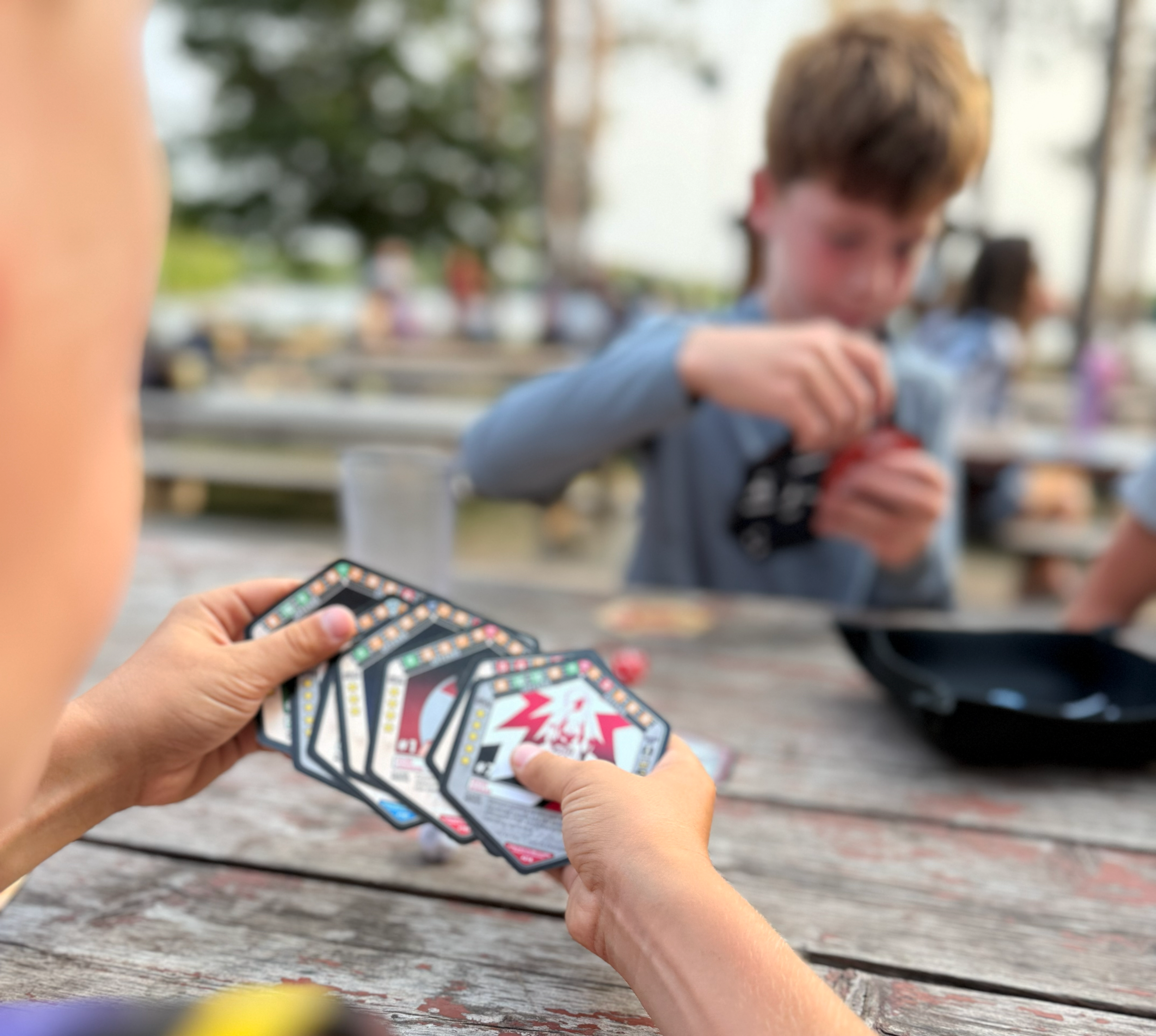 A person holding basketball playing cards while sitting at a wooden table outdoors, with a blurred background of people and trees.