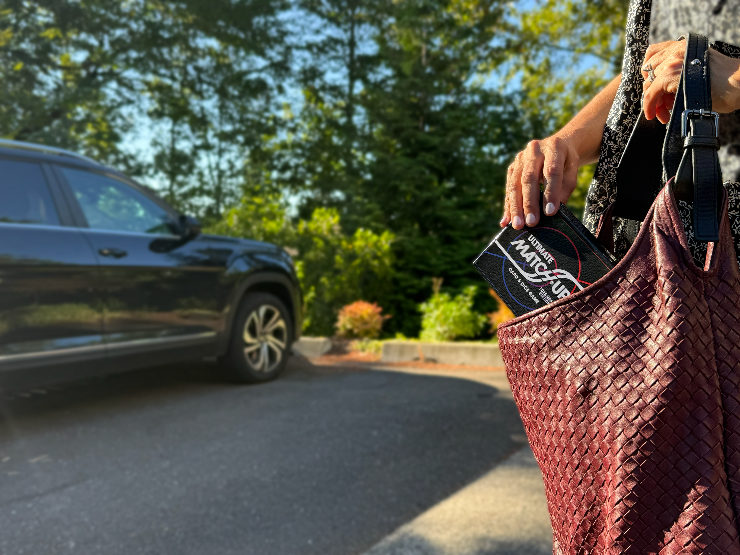 A person is holding a card game box labeled 'Ultimate Match' with a woven red bag over their arm, standing outdoors near cars and trees. Suggests portability of a game box.