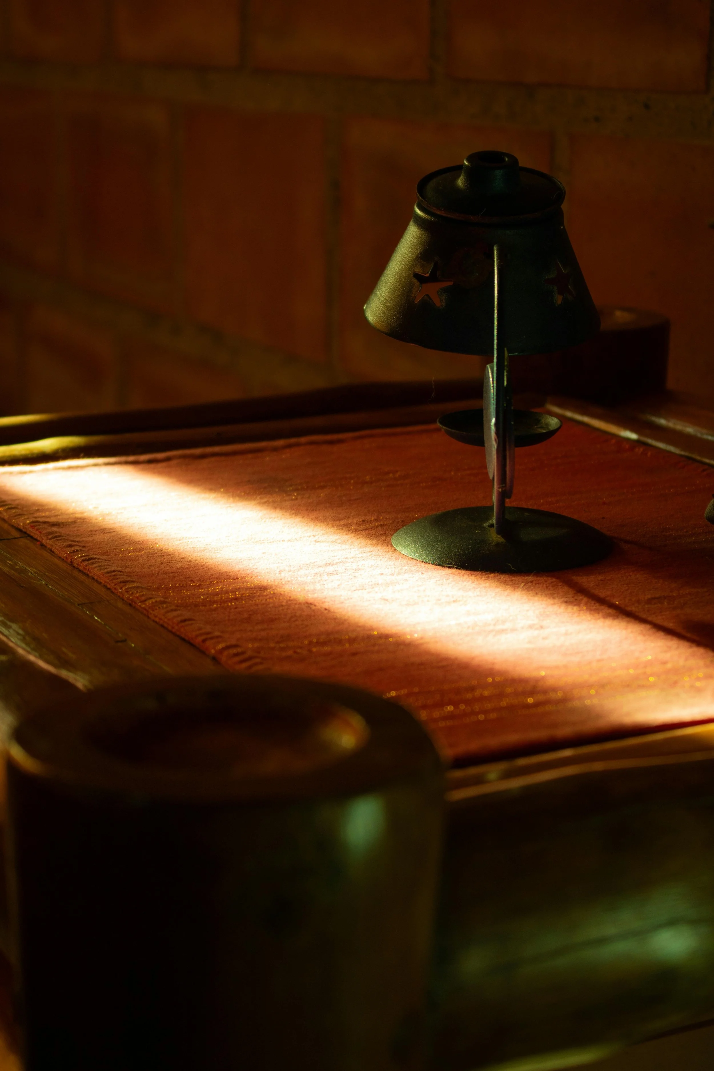 A small black metal lamp with star-shaped cutouts, placed on a wooden table with a reddish tablecloth. The lamp is turned off, and the background features a brick wall.