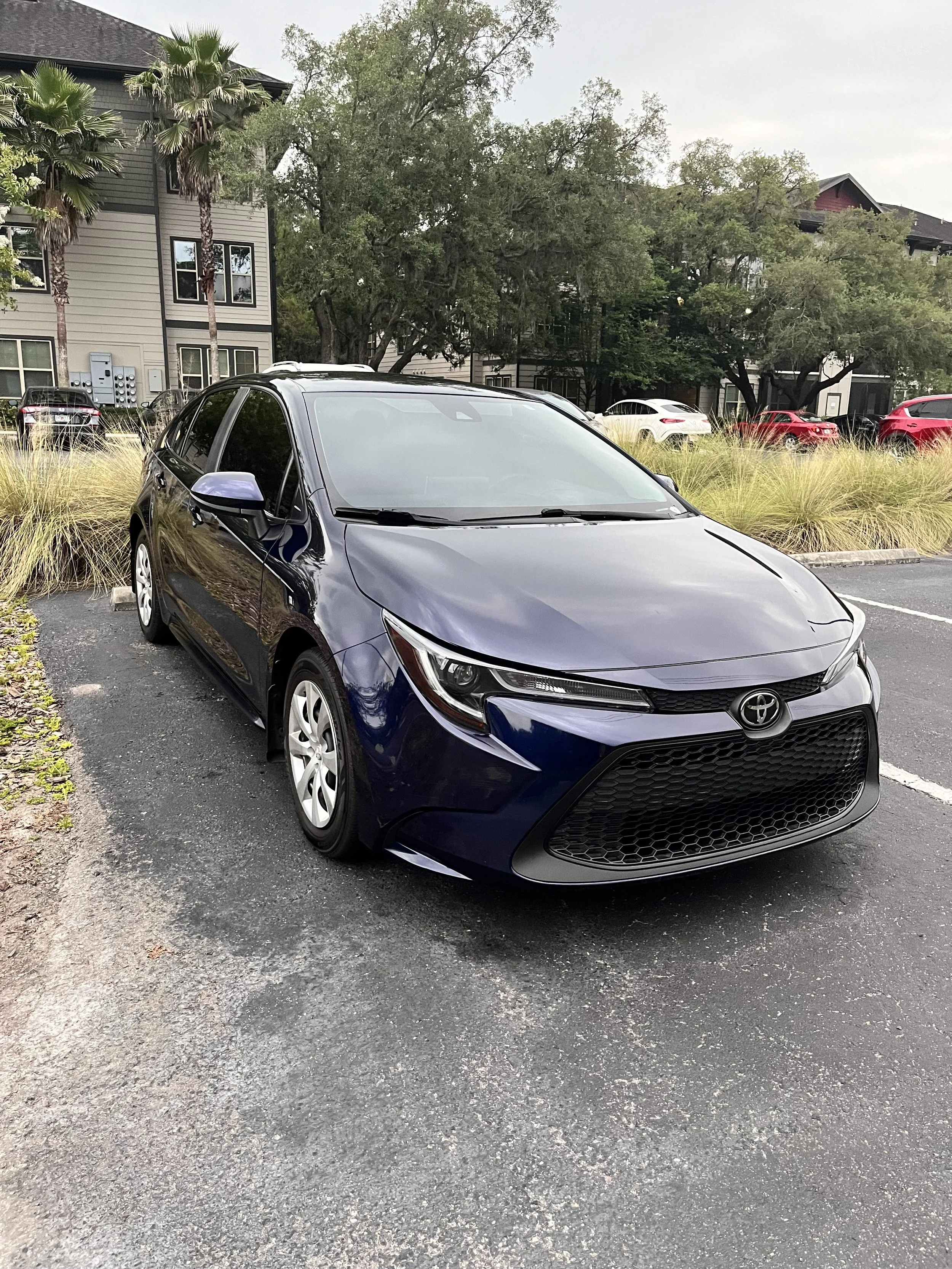A dark blue Toyota sedan parked in an outdoor parking lot with grassy landscaping and trees in the background.