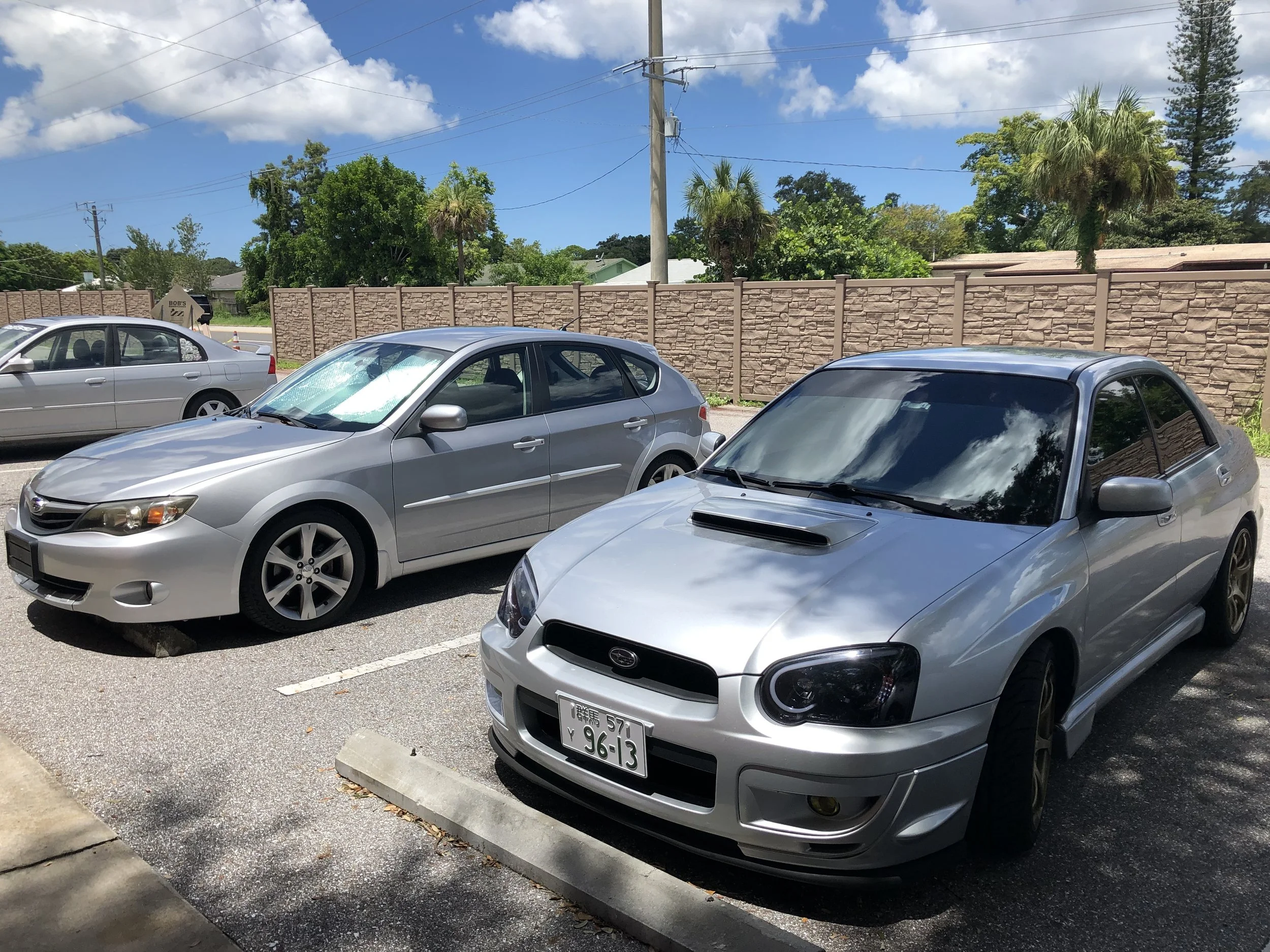 Two silver cars parked in a parking lot with a brick wall and trees in the background, under a partly cloudy sky.
