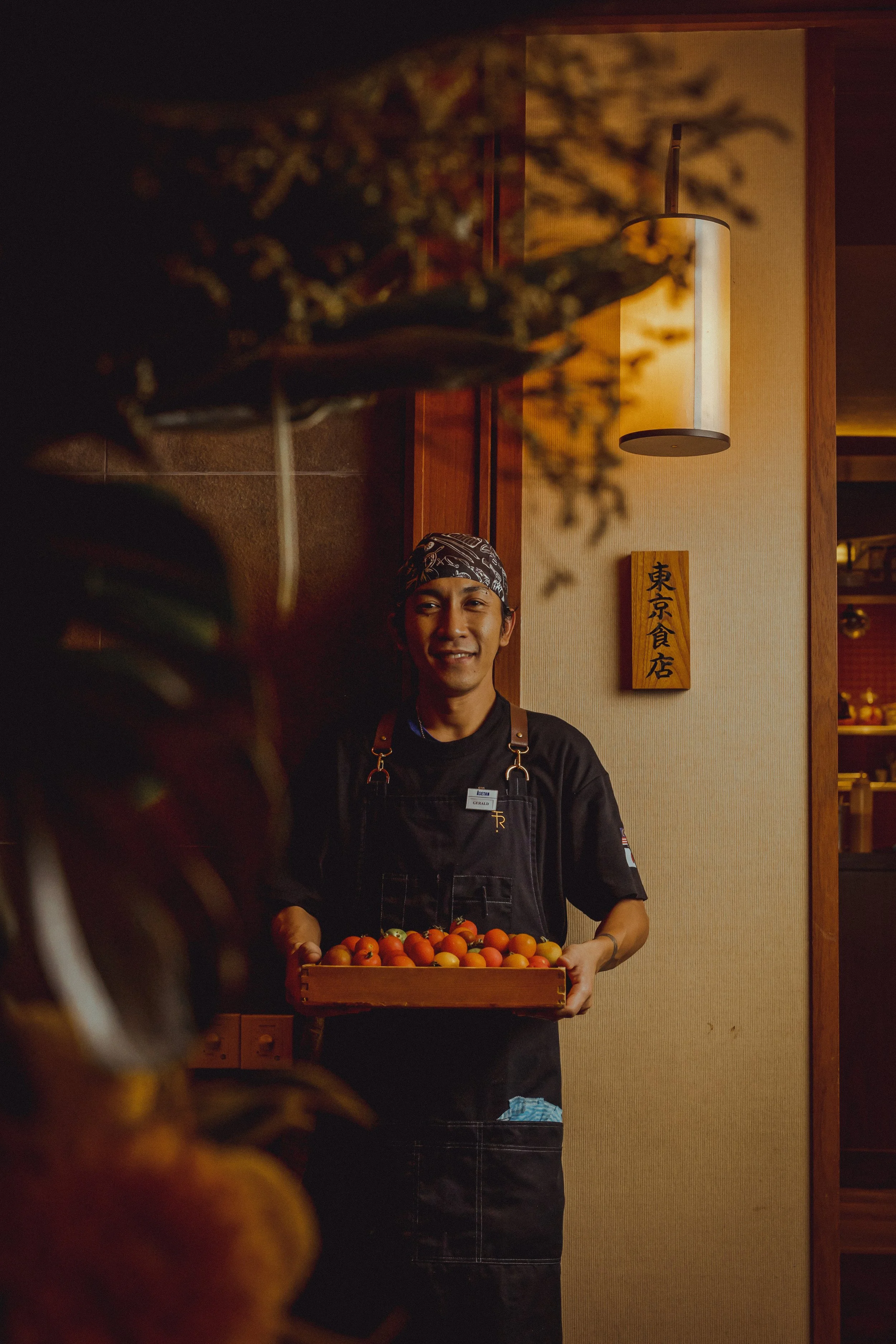 A chef holding a tray of cherry tomatoes, smiling, in a warmly lit restaurant with Japanese decor.