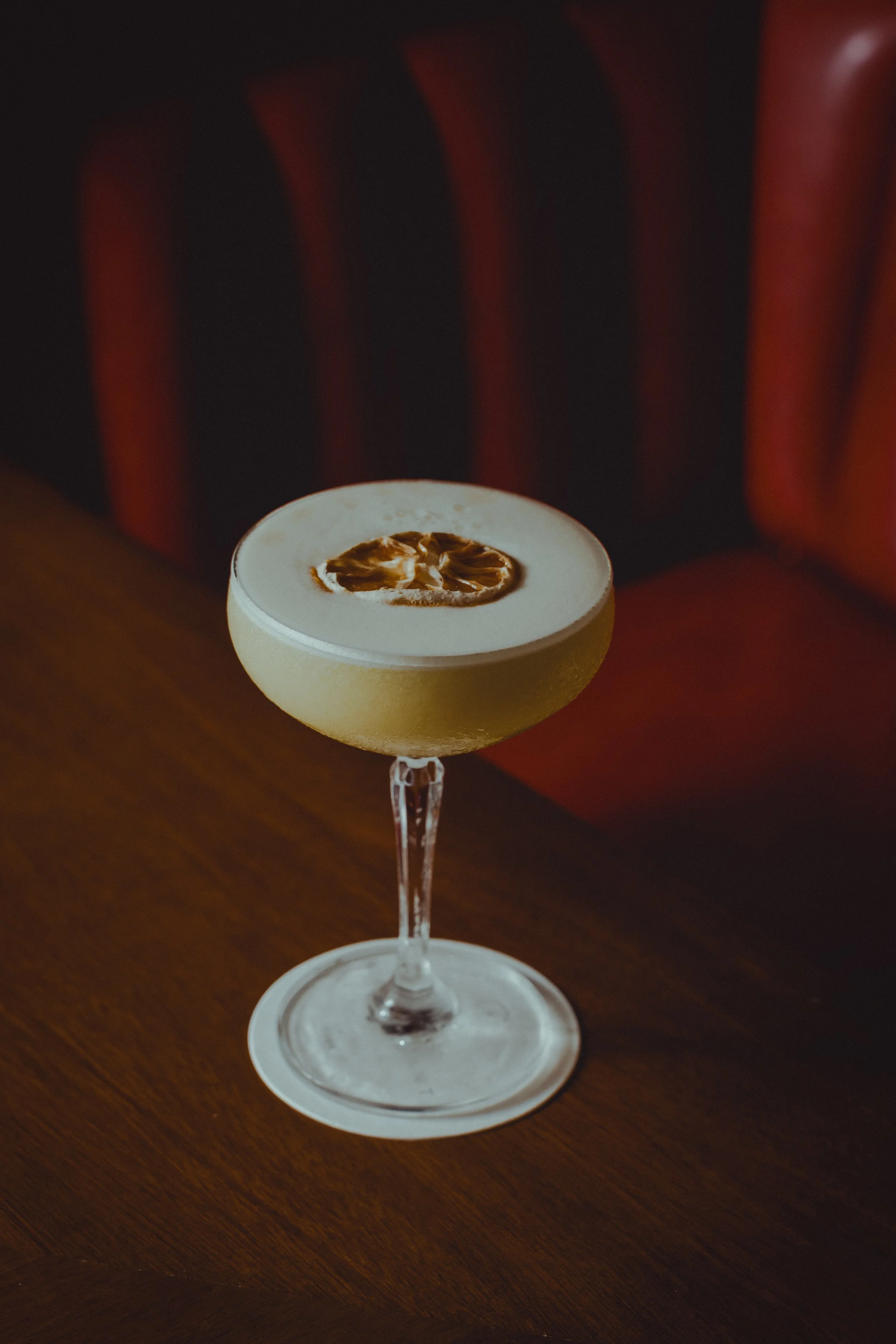 A cocktail drink in a cocktail glass with a lemon slice garnish on top, placed on a wooden table with a red and black striped upholstered background.