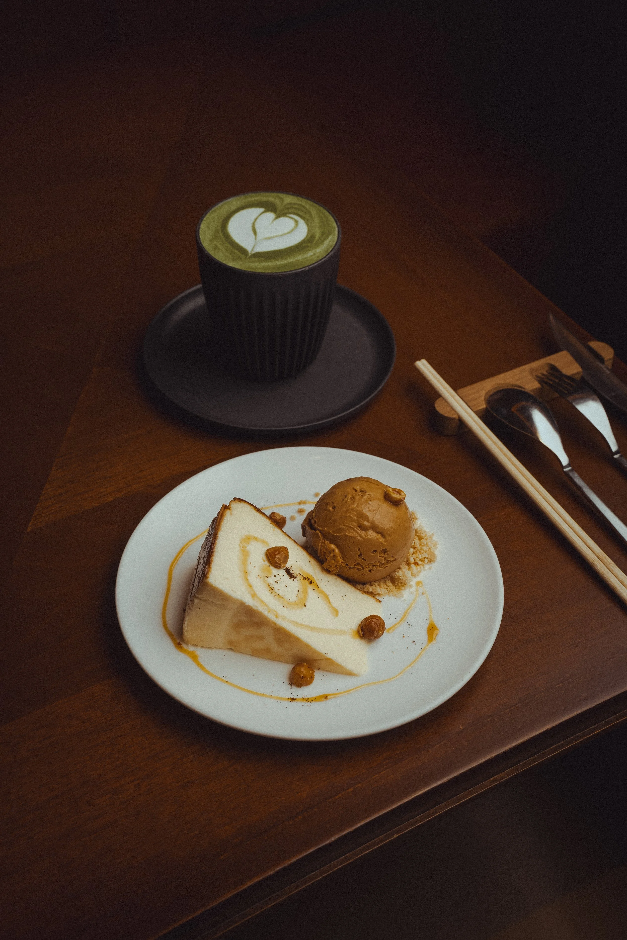 A dessert plate with a slice of cheesecake topped with caramel and a scoop of caramel ice cream, garnished with caramel drizzles and nuts, placed on a wooden table alongside a cup of matcha latte with latte art, chopsticks, and utensils.