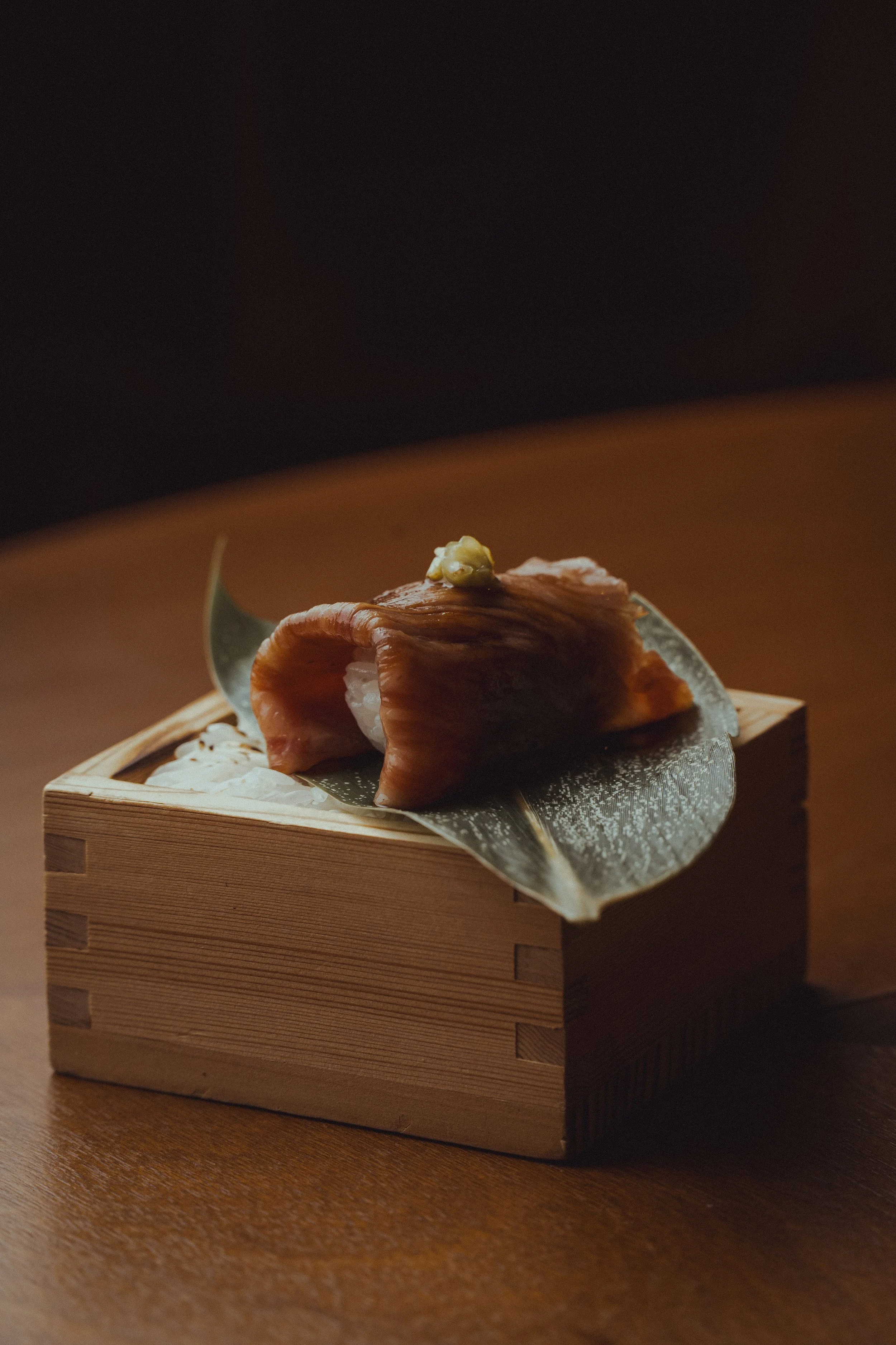 A piece of sushi with fish and wasabi on top, placed on a piece of leaf on a small wooden box, with a dark background.