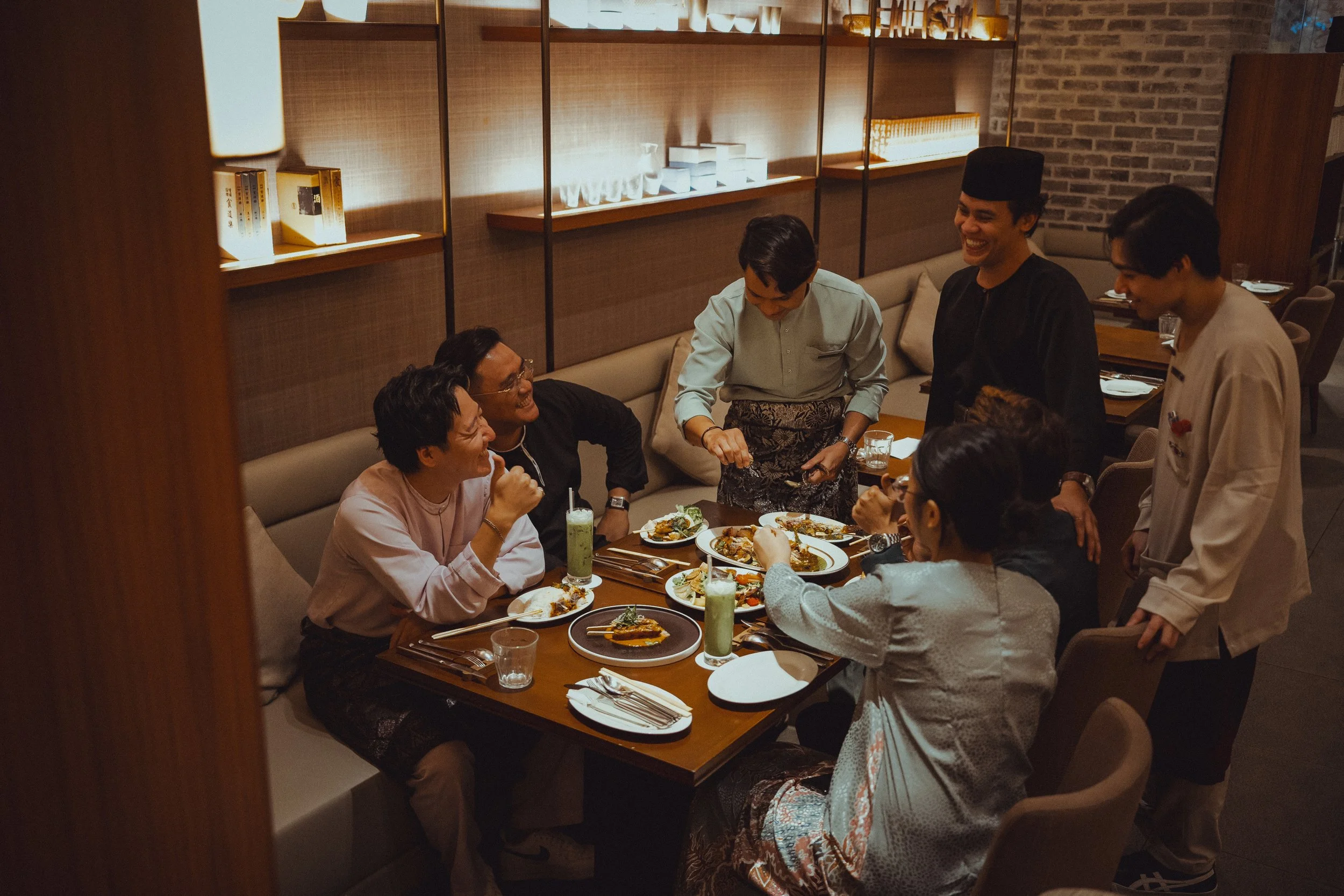 A group of people enjoying a meal together in a restaurant, with some standing and others sitting around a table filled with various dishes and drinks, smiling and engaging in conversation.