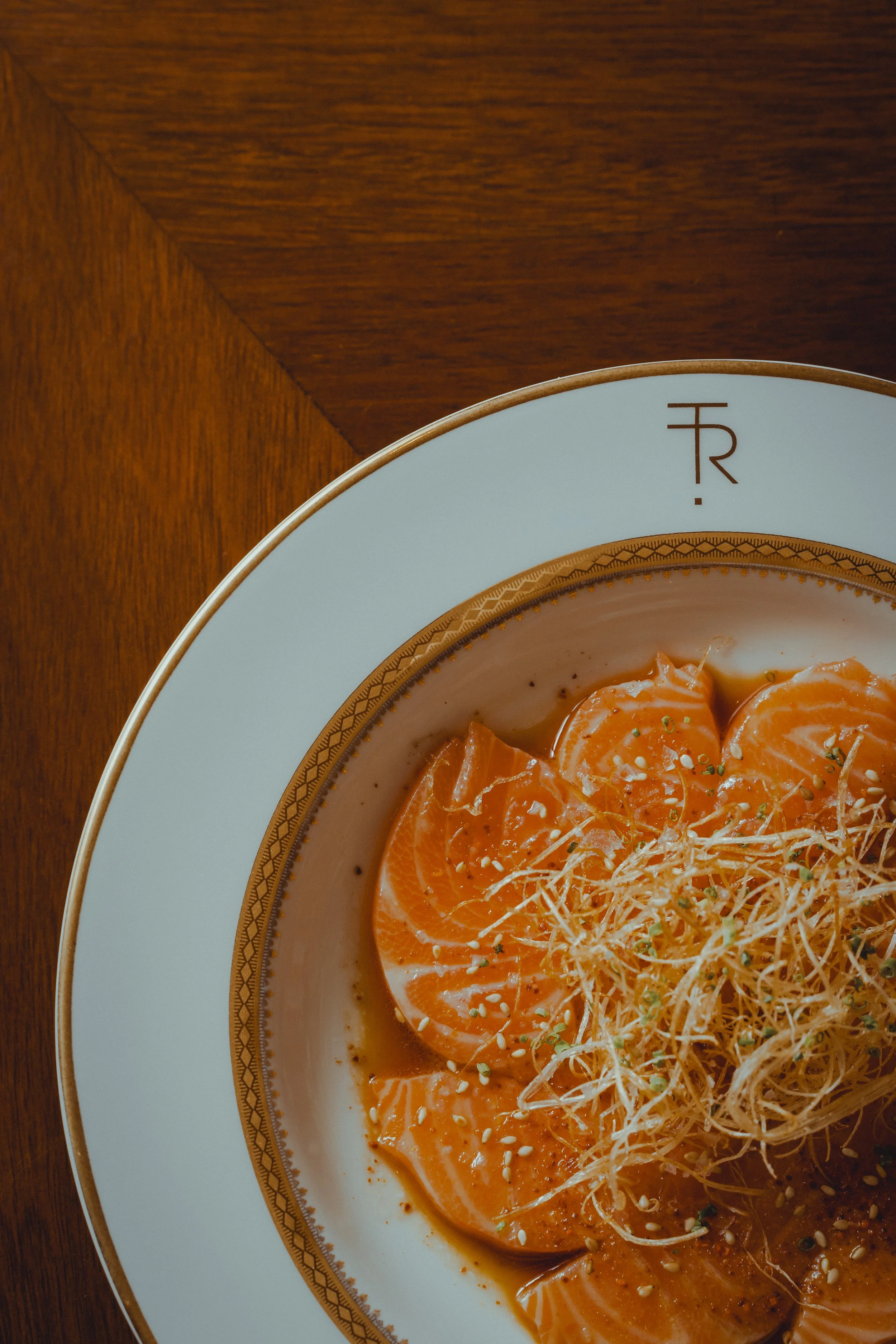 Close-up of a plate of sliced salmon garnished with sprouts, sesame seeds, and herbs, on a decorative white and gold-rimmed plate, placed on a wooden surface.