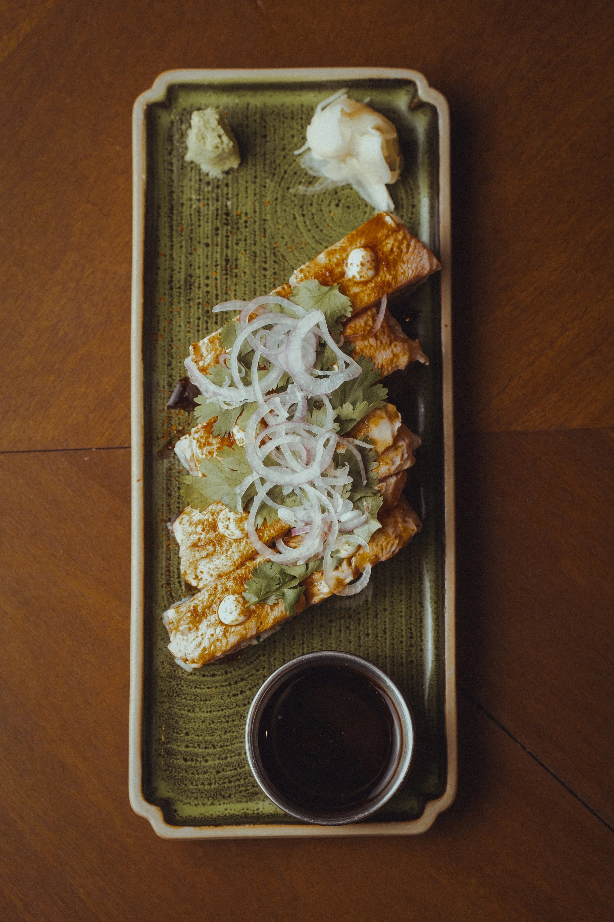 Sushi rolls topped with sliced onions, cilantro, and a drizzle of sauce, accompanied by wasabi, ginger, and soy sauce in a rectangular green dish on a wooden table.