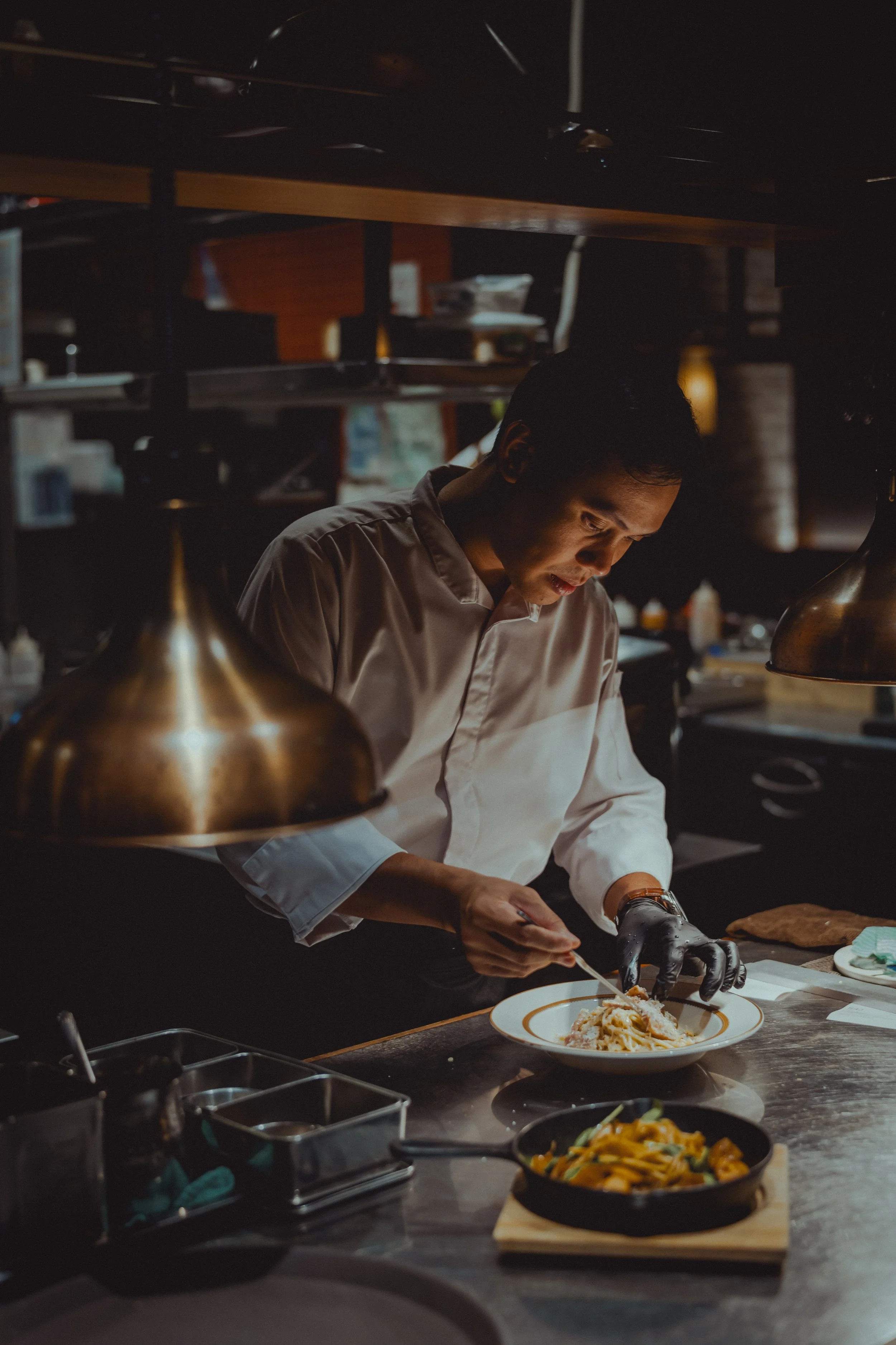 A chef in a professional kitchen garnishing a plate of pasta. The kitchen has warm lighting, with hanging lamps and shelves in the background.