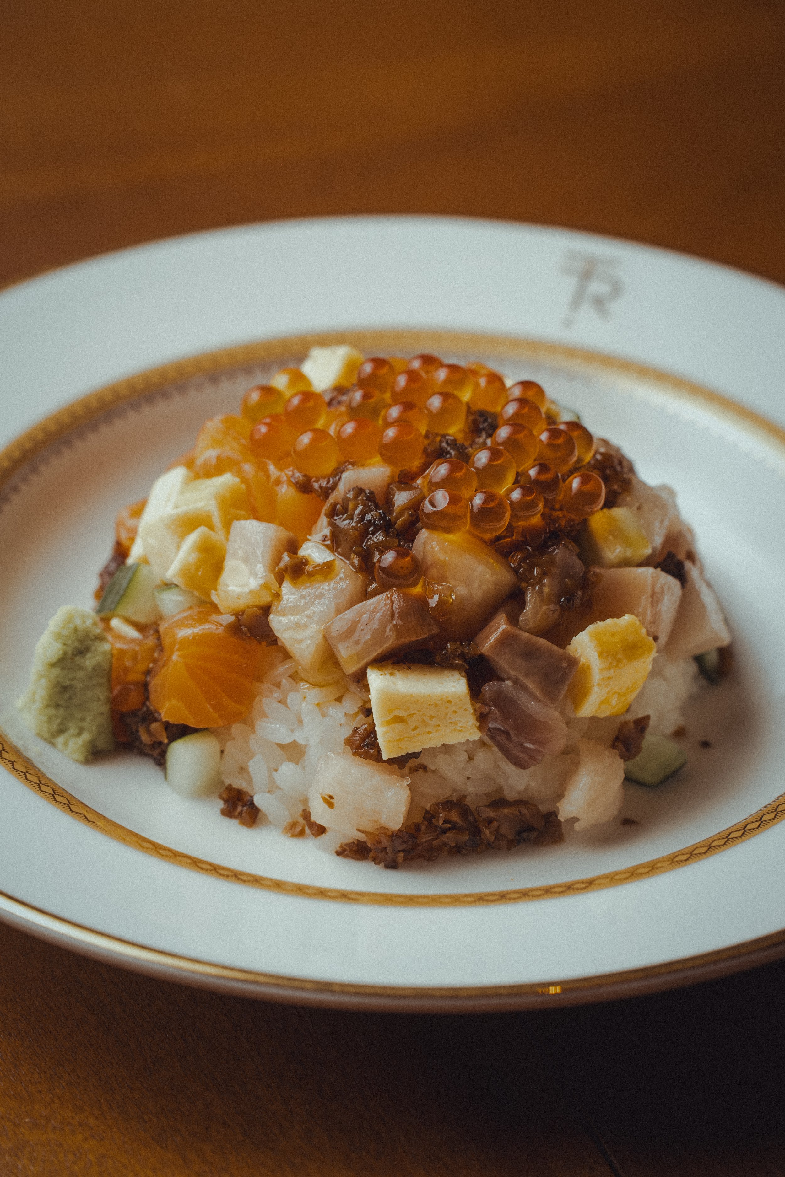 A plate of sushi topped with orange fish roe and various diced ingredients, served on a white and gold-rimmed plate.