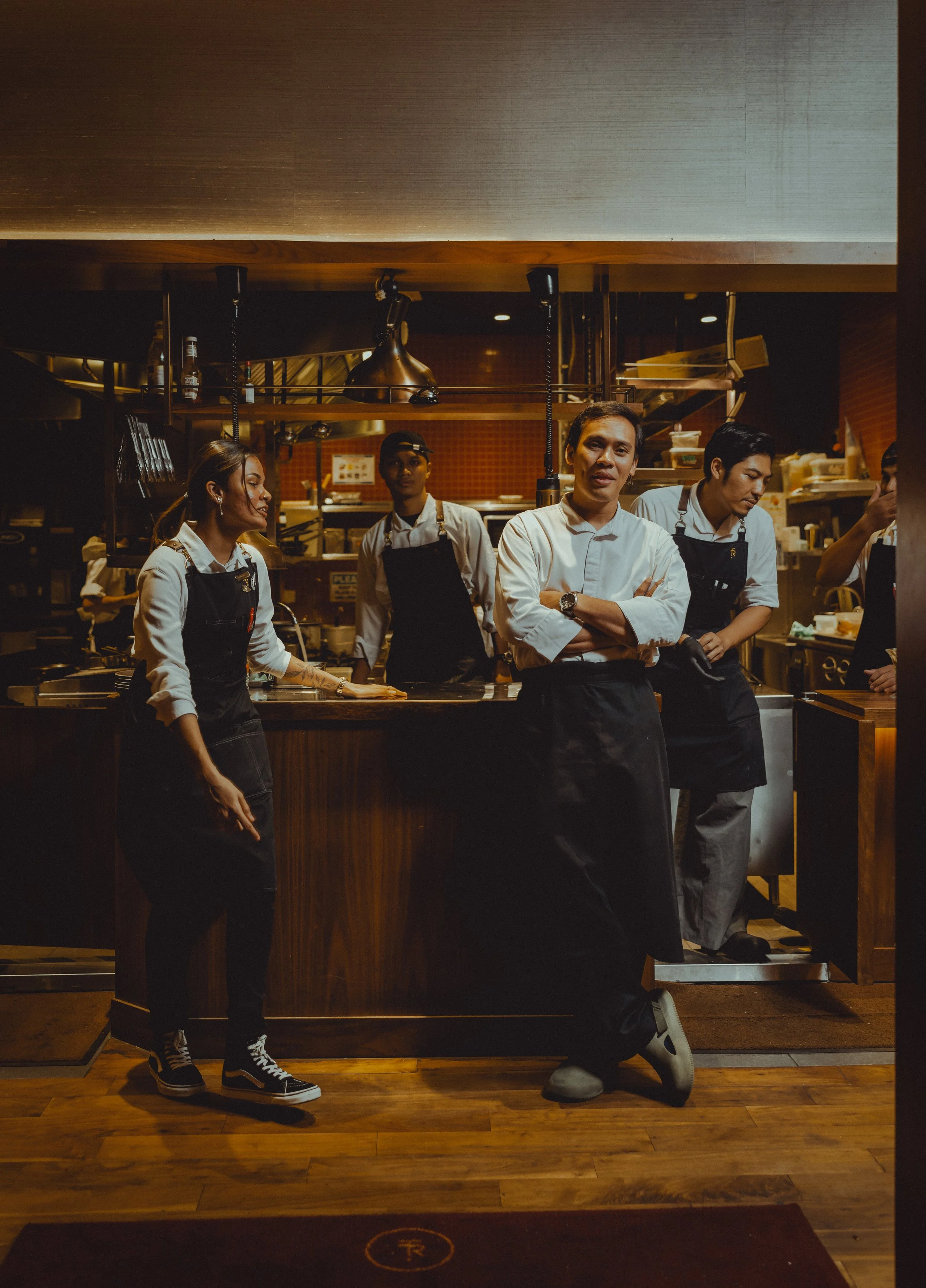 Group of restaurant staff in kitchen, including a man with arms crossed and three women in uniform, working behind the counter.