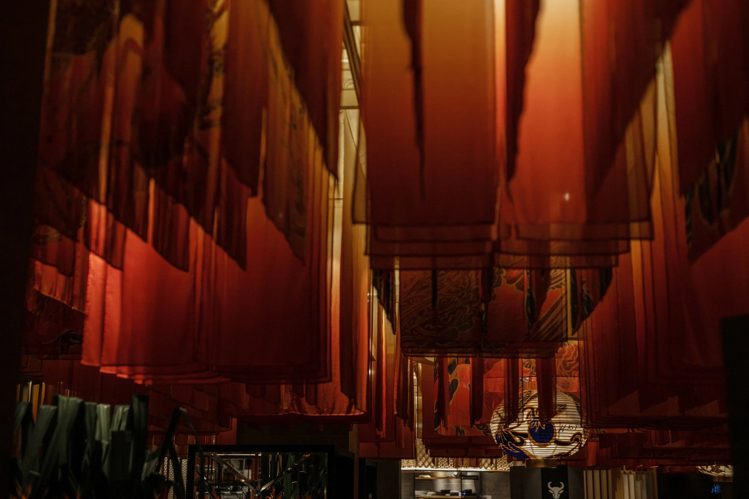 Interior of a dimly lit space with hanging paper lanterns and fabric banners with traditional Japanese patterns.