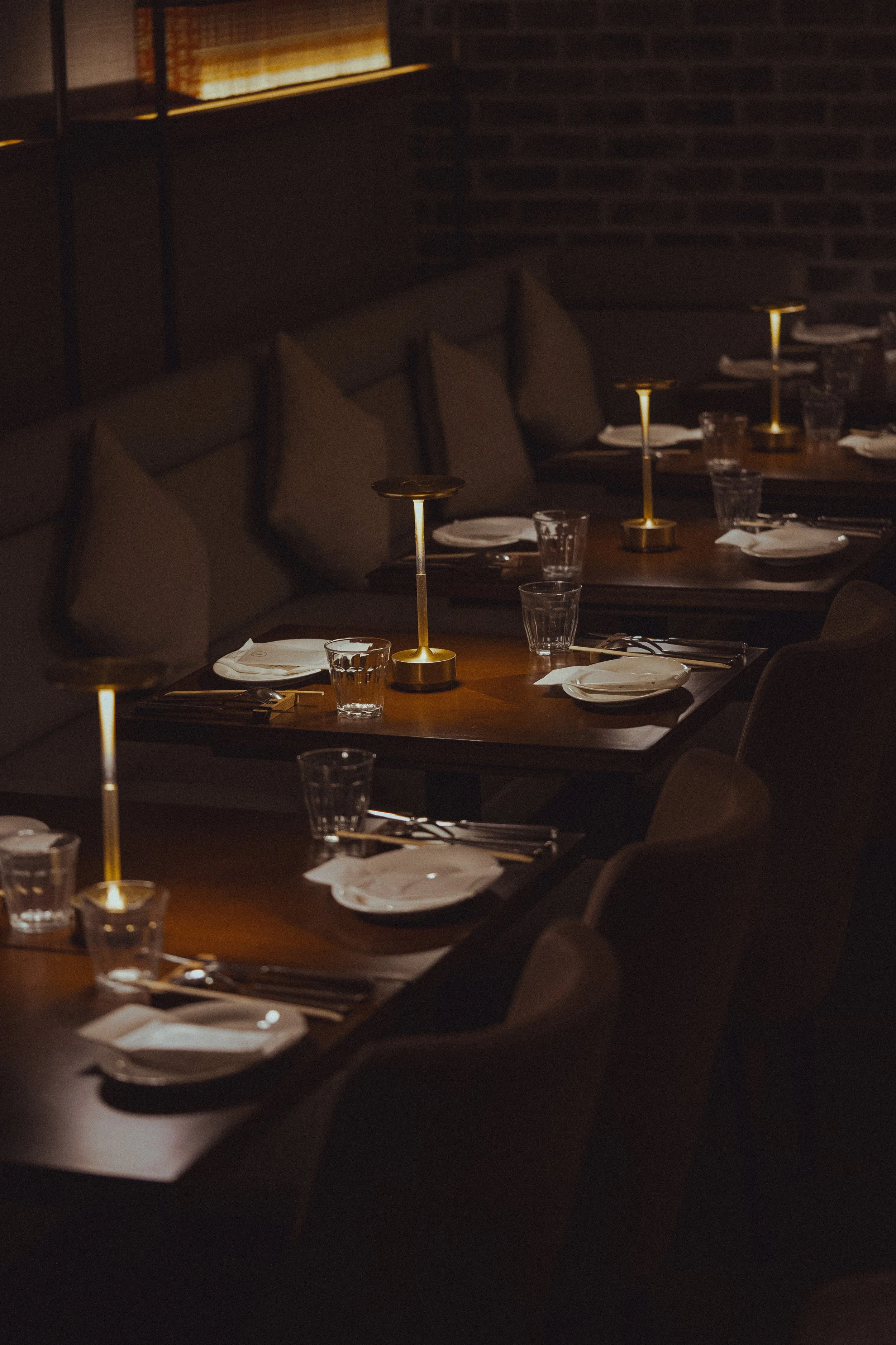 A dimly lit restaurant with neatly arranged tables, each set with white plates, silverware, water glasses, and golden candle holders.