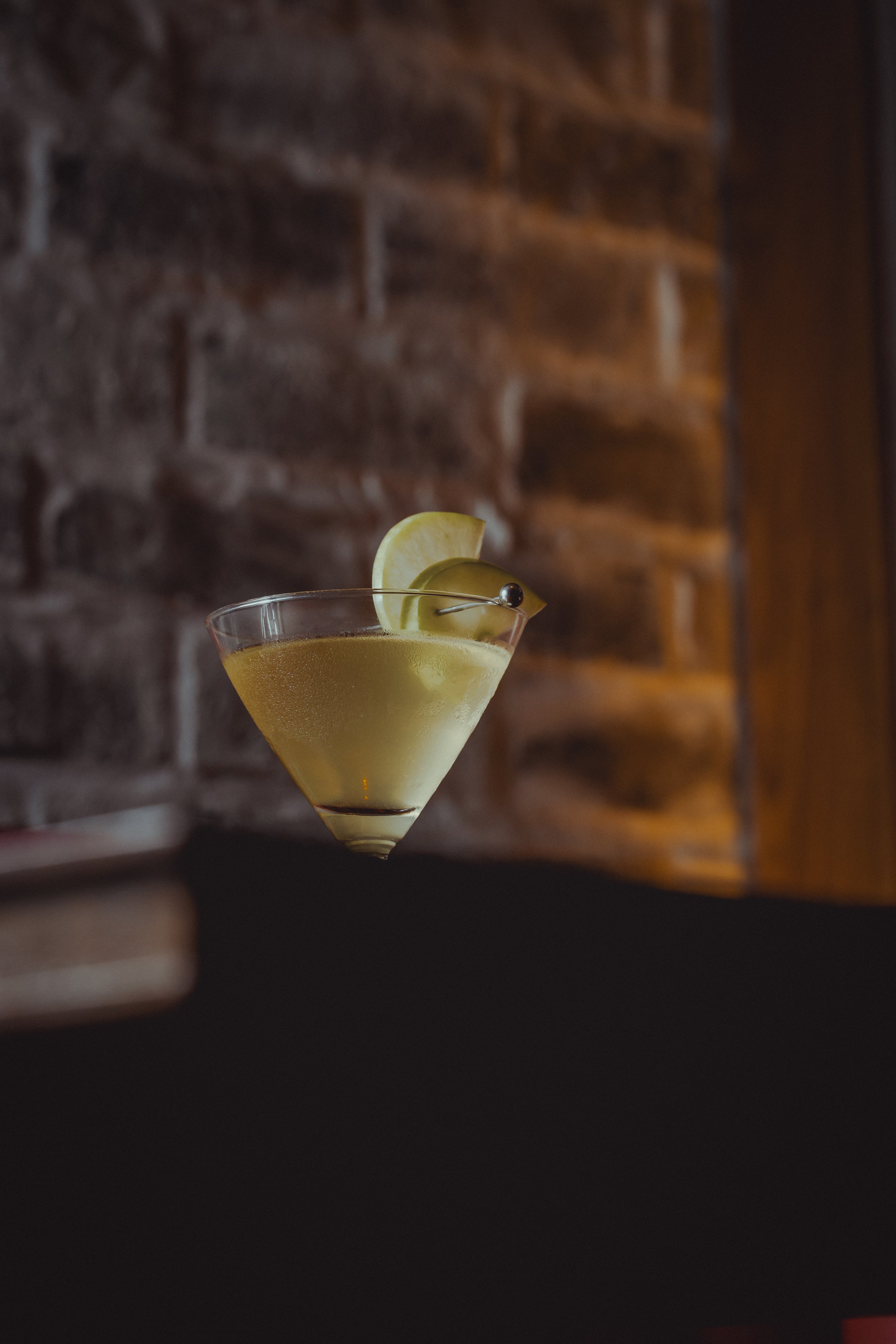 A cocktail glass filled with a yellow-green drink, garnished with a lemon wedge and a green olive on a cocktail pick, against a blurred brick wall background.