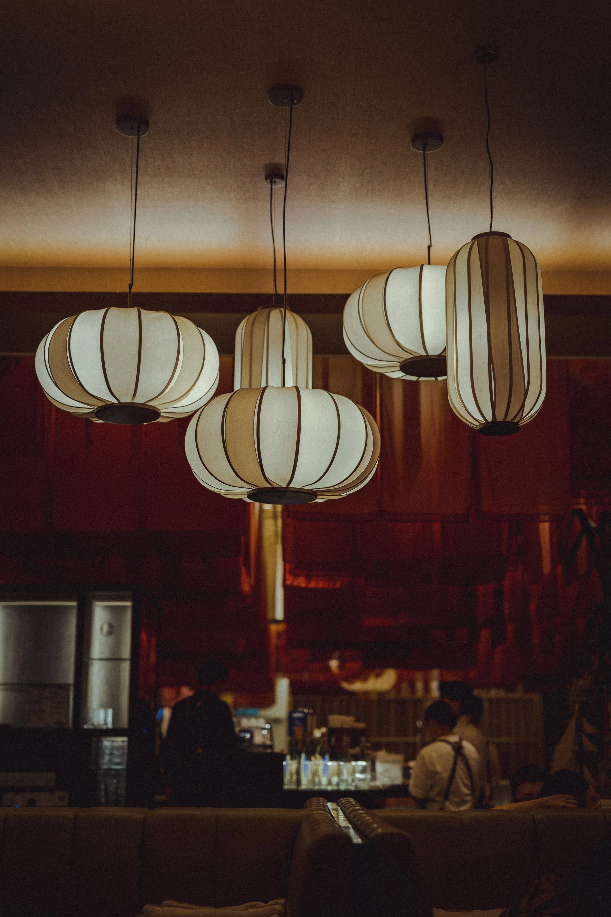Pendant lamps hanging from a ceiling in a dimly lit restaurant or cafe, with people working or socializing in the background.