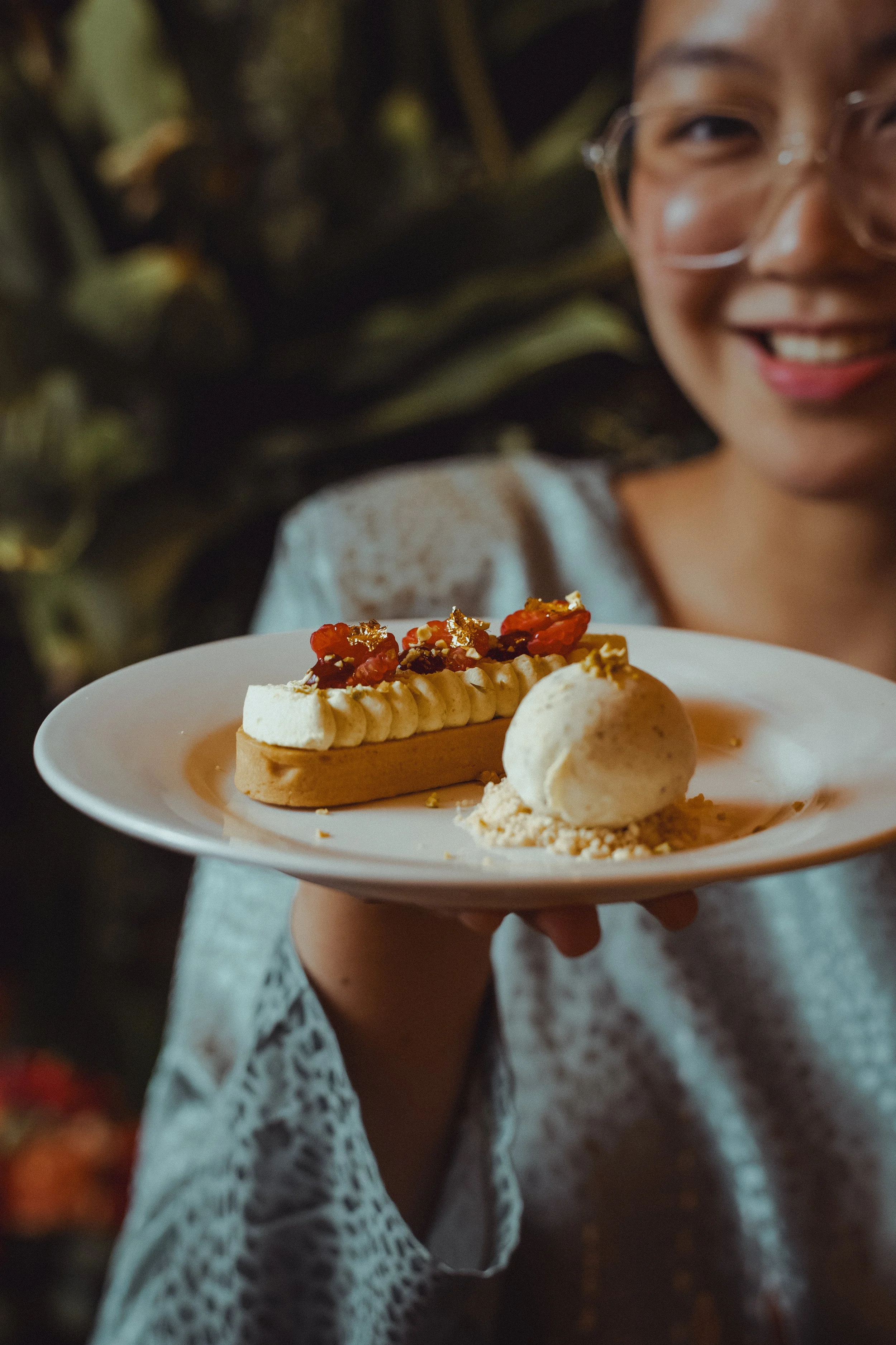 A woman smiling while holding a plate with a dessert featuring a rectangular pastry topped with whipped cream, dried red fruits, and gold leaf, alongside a scoop of vanilla ice cream.