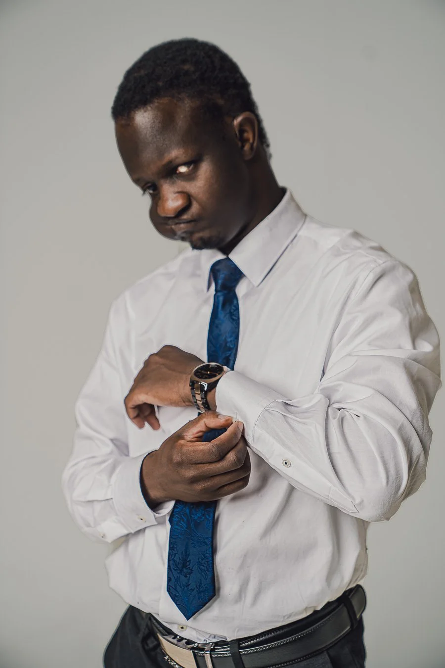 A man in a white dress shirt and blue tie adjusting his cuff, wearing a watch, with a serious expression, against a plain gray background.