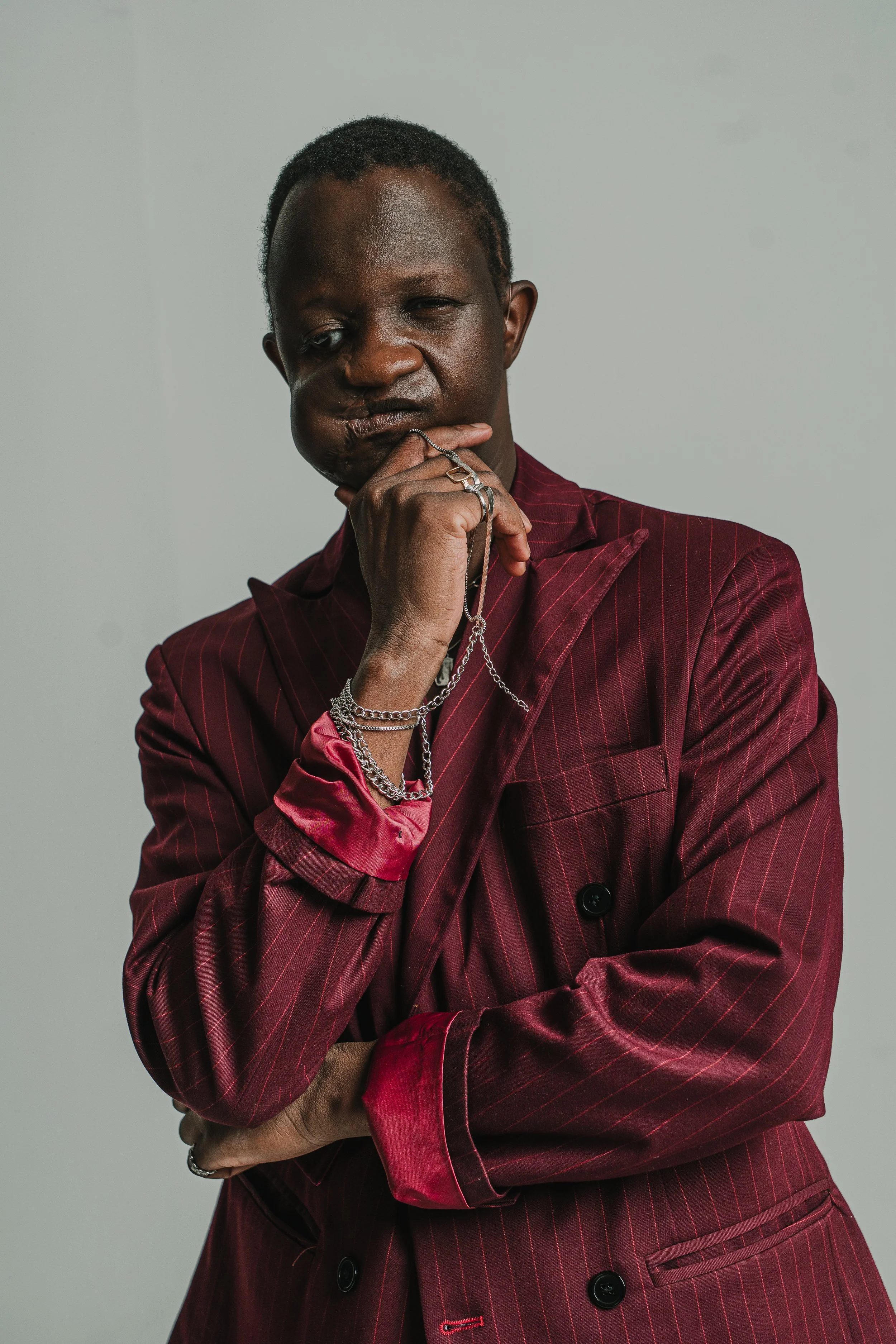A person with dark skin and short hair wearing a red pinstripe blazer with satin pink cuffs, posed with one hand on their chin, adorned with various silver rings and bracelets, against a plain gray background.