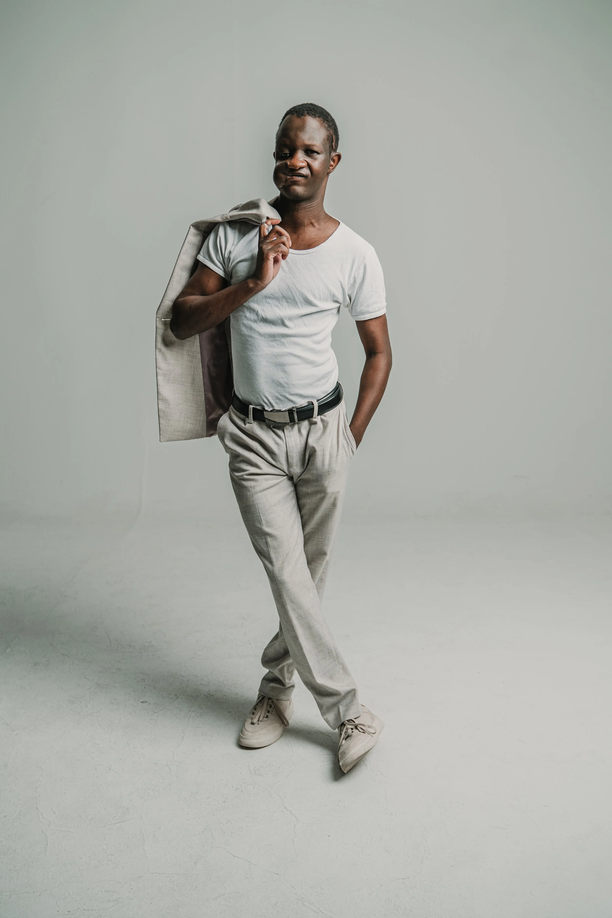 A person with dark skin and short hair wearing a white t-shirt, beige pants, and beige sneakers poses with a casual stance against a plain light grey background. They carry a beige jacket over their shoulder and have one hand in their pocket.