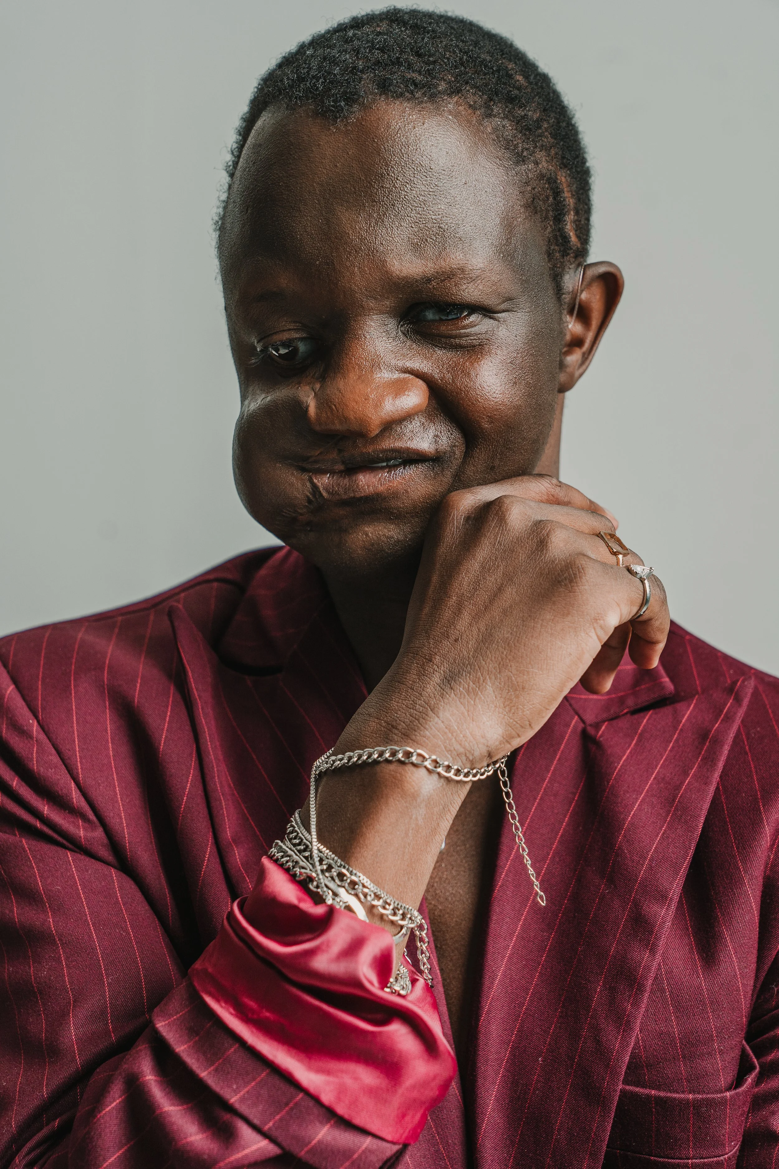 A person with dark skin and short curly hair posing with their hand near their face. They are wearing a maroon pinstripe suit, silver jewelry, and a shiny red satin shirt.