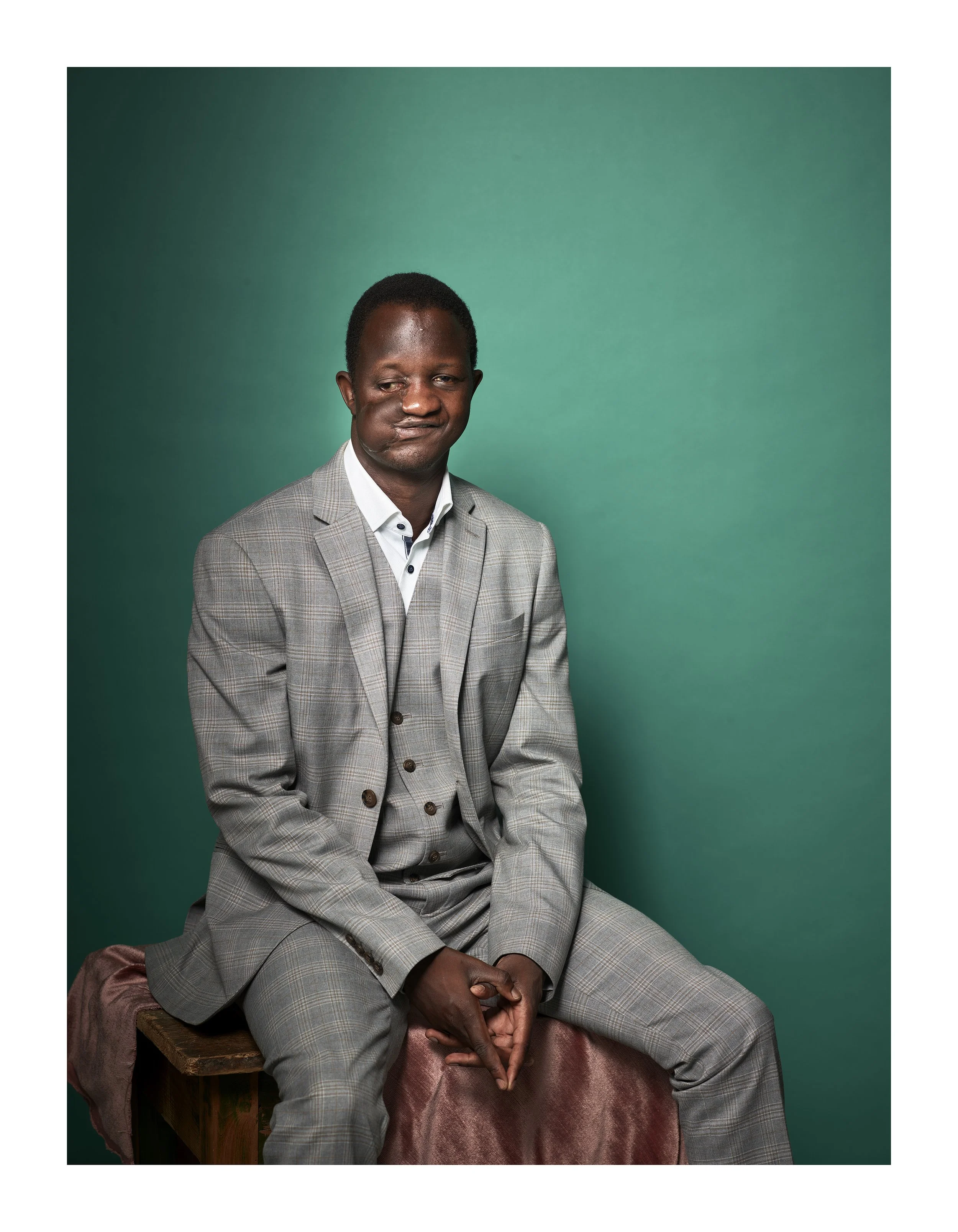 Fidel Strub in a gray plaid suit sitting on a wooden bench against a solid teal background, looking slightly to the side with a serious expression.