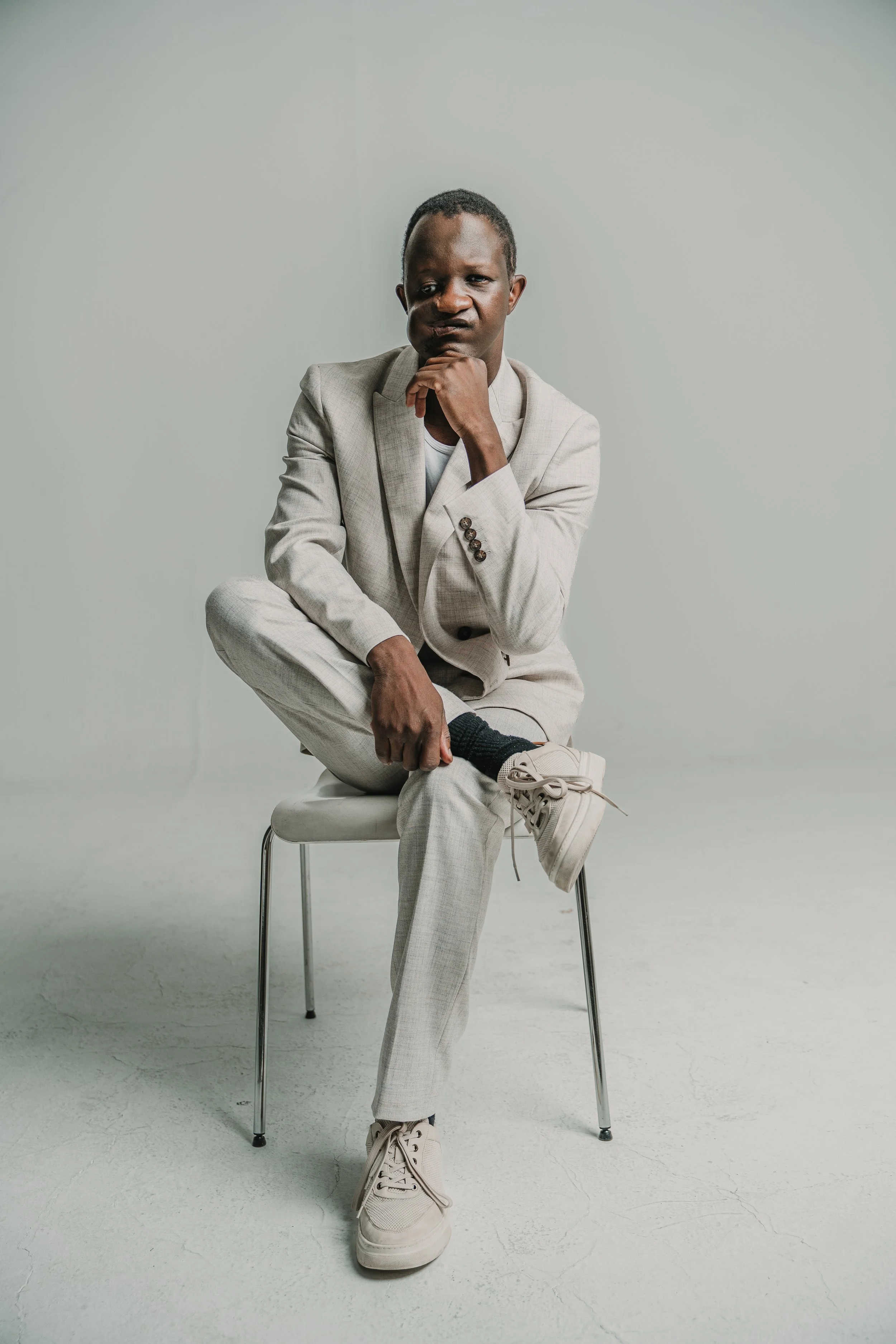 A man dressed in a light-colored suit is sitting on a white chair against a plain light background, with one leg crossed over the other and his chin resting on his hand, gazing thoughtfully.