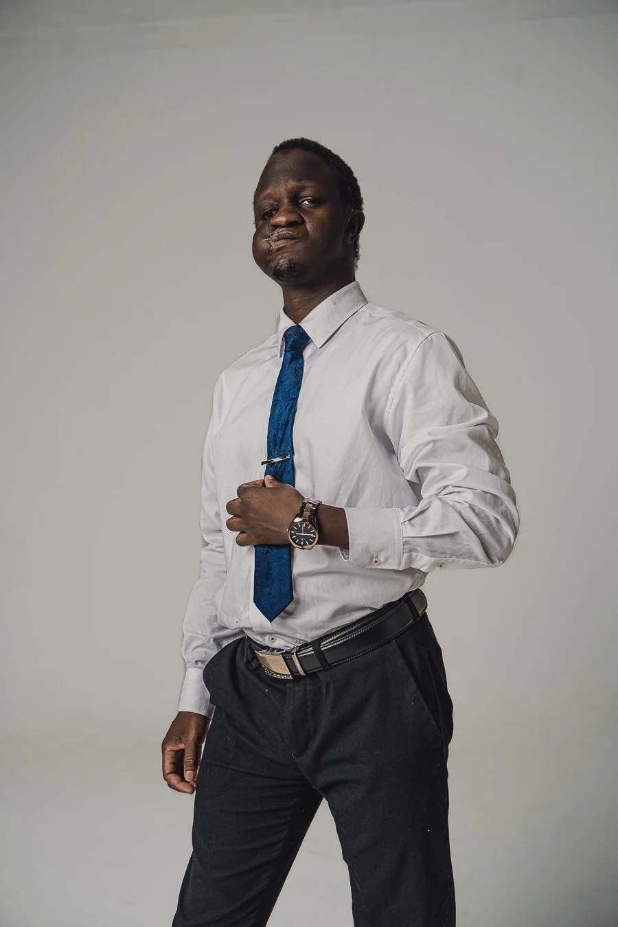 A man wearing a white dress shirt, blue tie, and black pants, standing against a plain background, holding his tie with one hand, and wearing a wristwatch.