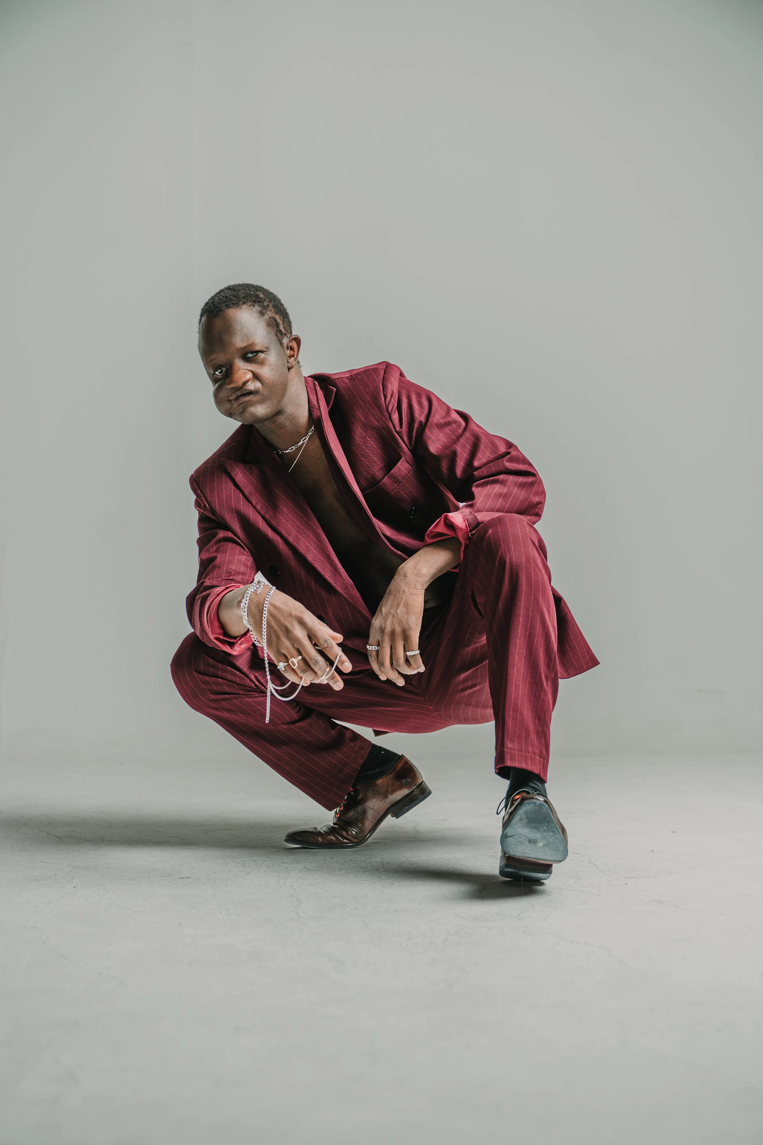 A person with dark skin and short hair squat down, wearing a maroon pinstripe suit and brown shoes, with accessories including rings, necklaces, and bracelets, set against a plain light grey background.