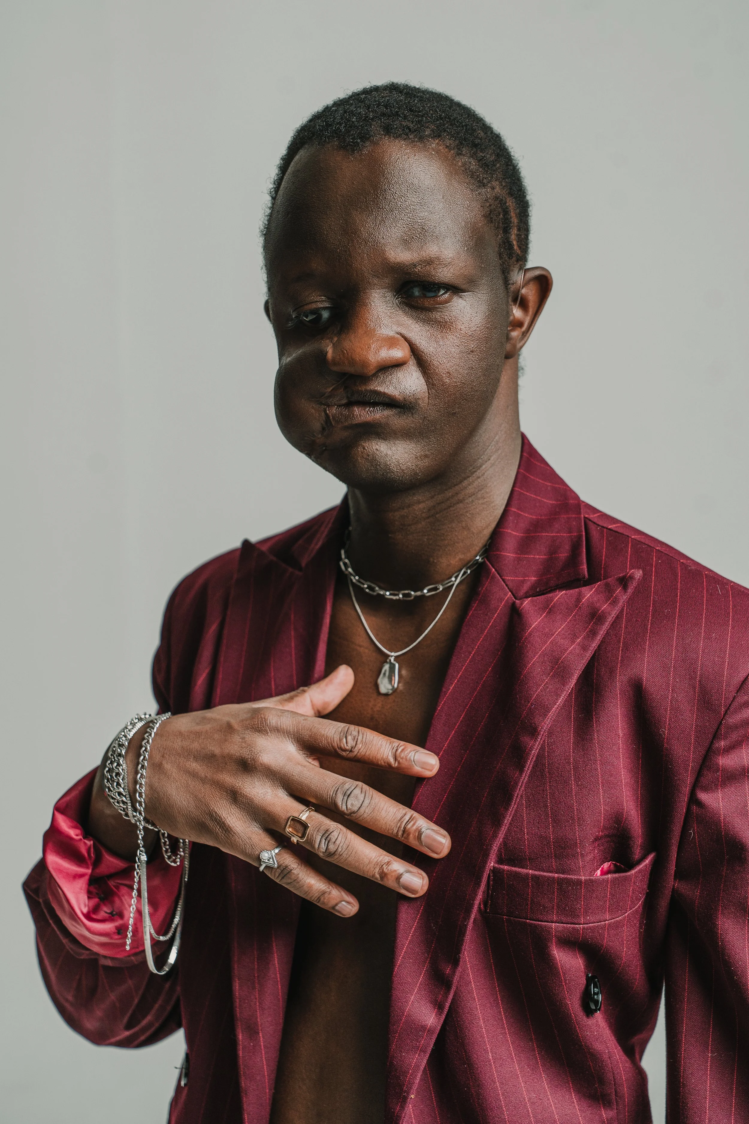 A person with dark skin wearing a red pinstripe suit jacket, accessorized with multiple silver necklaces, bracelets, and rings. They have short curly hair and are making a gesture with their fingers, touching their chest with one hand. The background