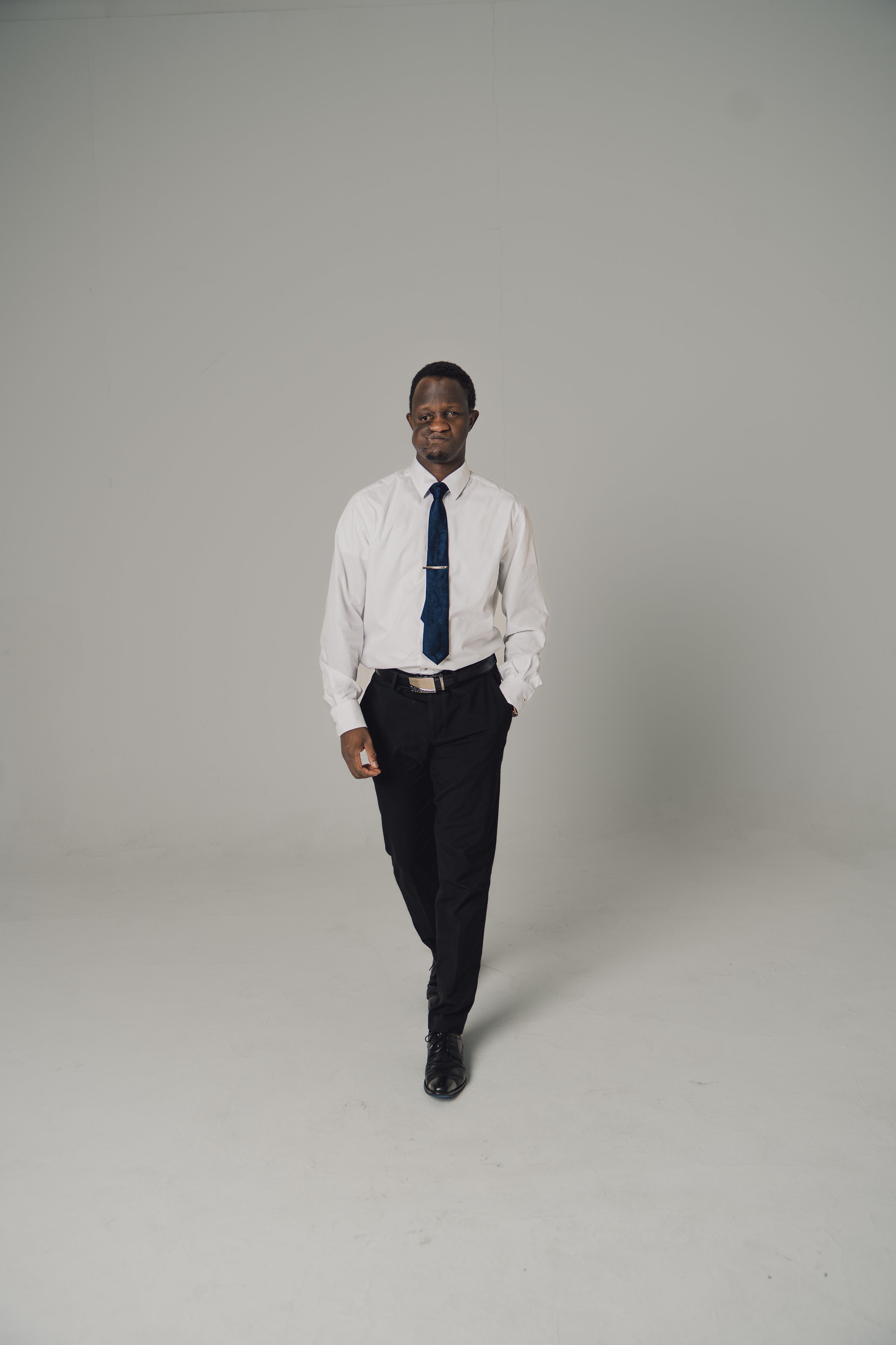 A man dressed in a white shirt, black pants, and black shoes walking with one hand in his pocket against a plain gray background. He is wearing a dark blue tie with a tie clip.
